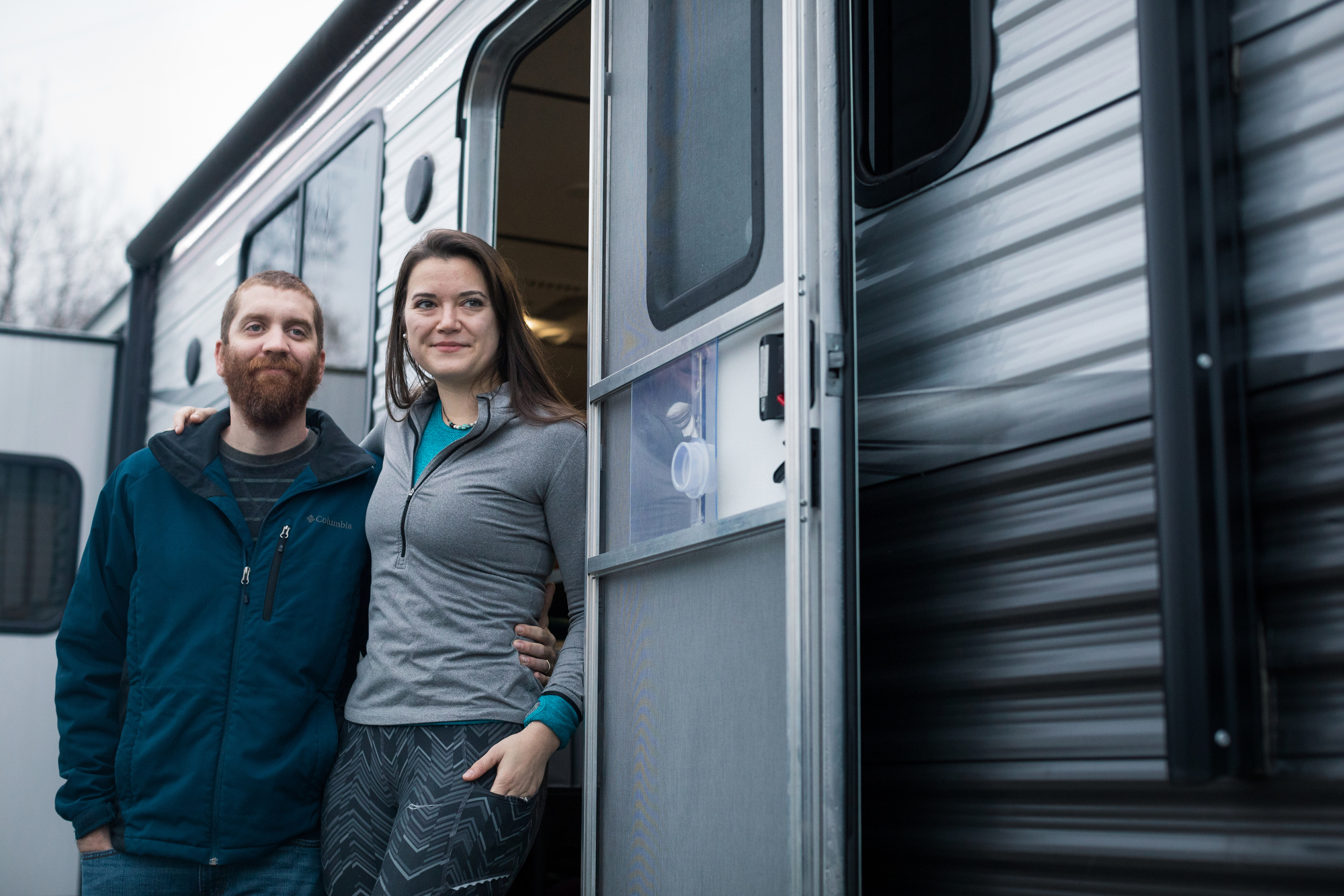 A man and woman stand just outside the door of a silver and gray RV.