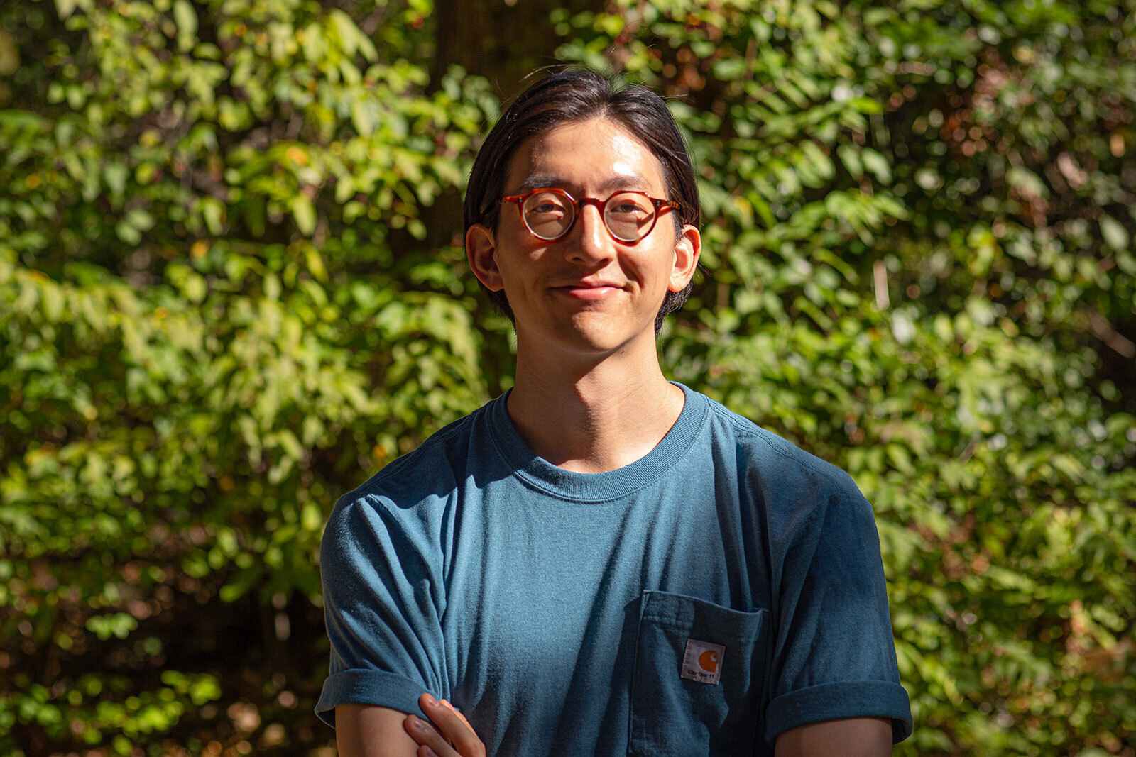 An image of a man smiling for a photo outside in front of a large green shrub.