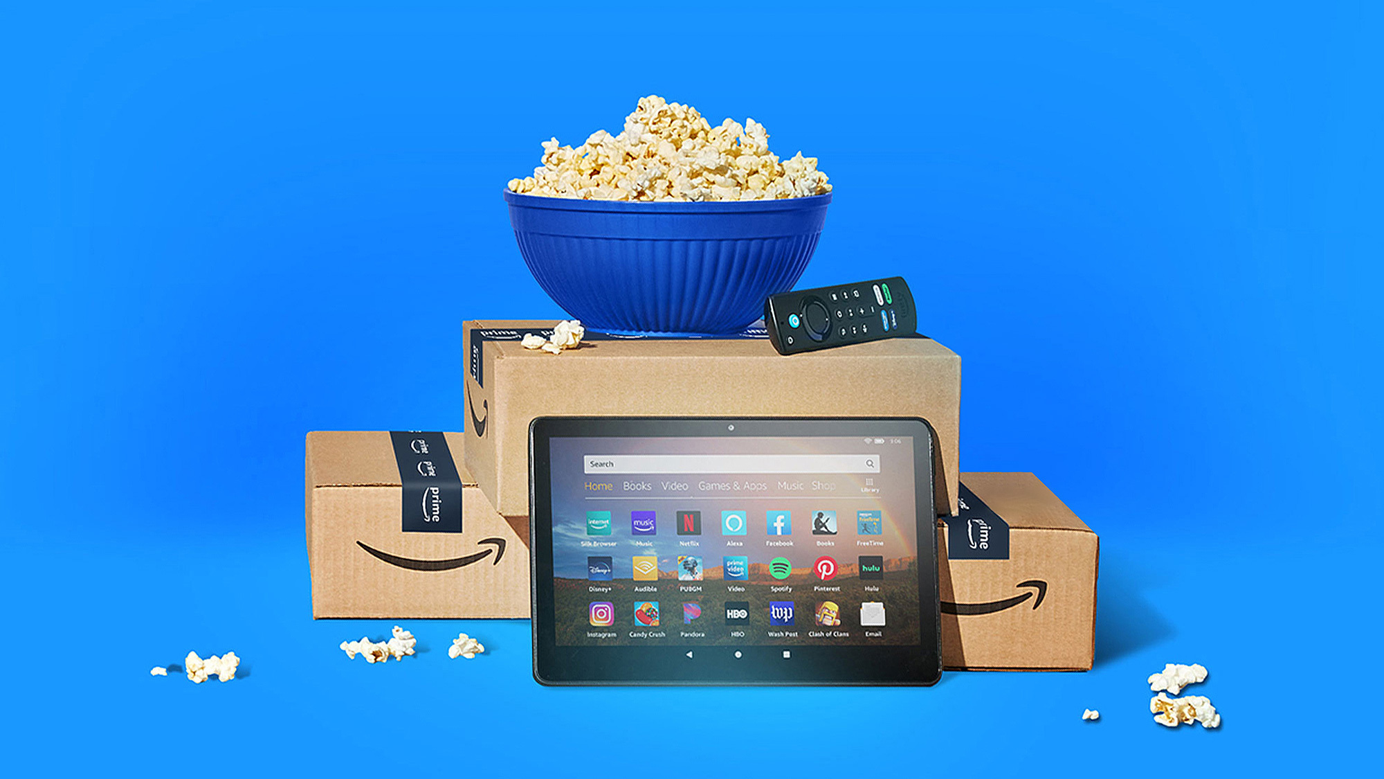 Das Bild zeigt eine Schüssel Popcorn, die auf einem Stapel von Amazon-Kartons steht. Vor den Schachteln liegen ein Tablet und Fernbedienungen für den Fire TV. 