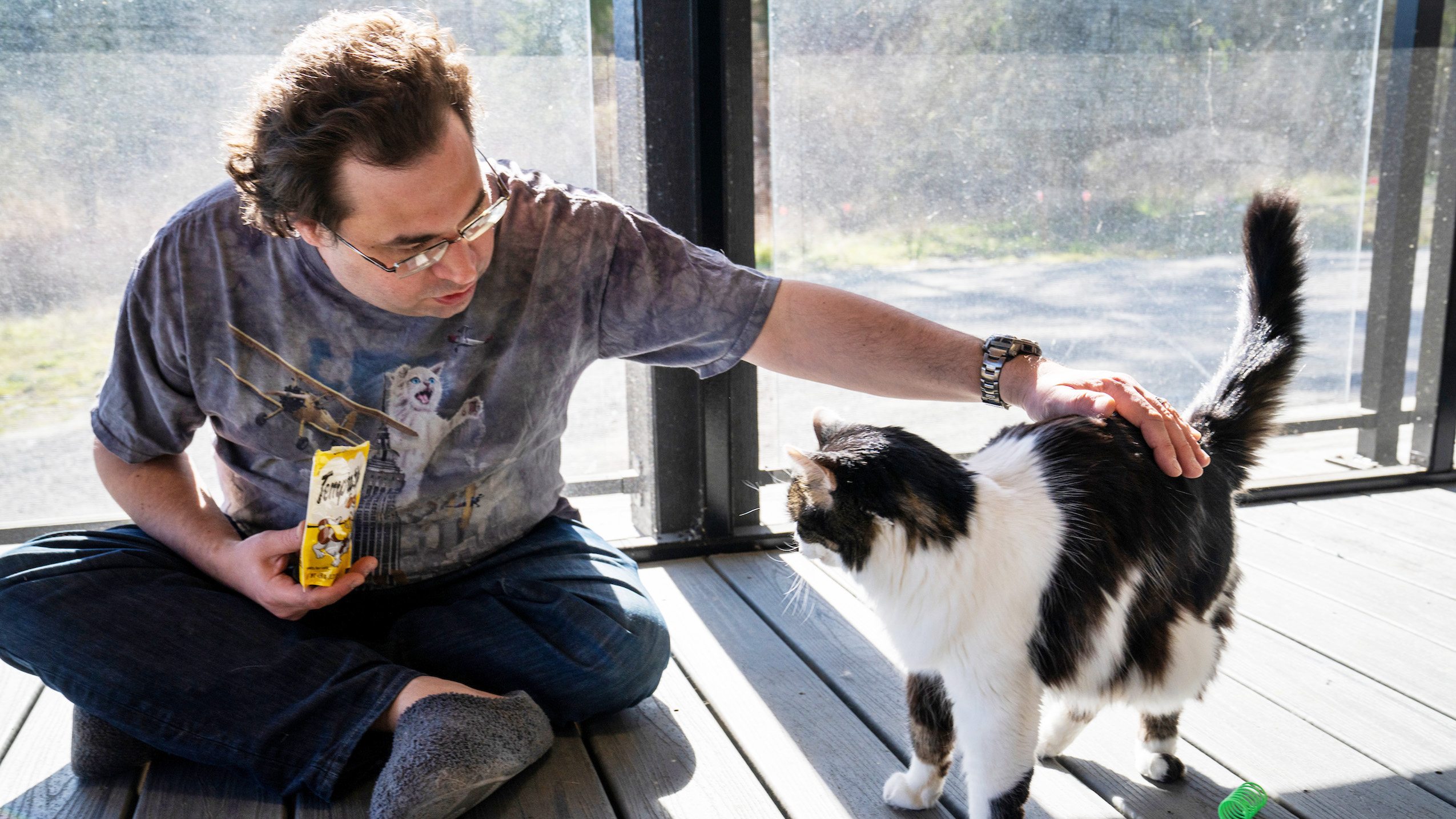 A photo of AWS VP and Distinguished Engineer Tom Scholl sitting on the floor at his home, holding a bag of cat treats and petting one of his cats.