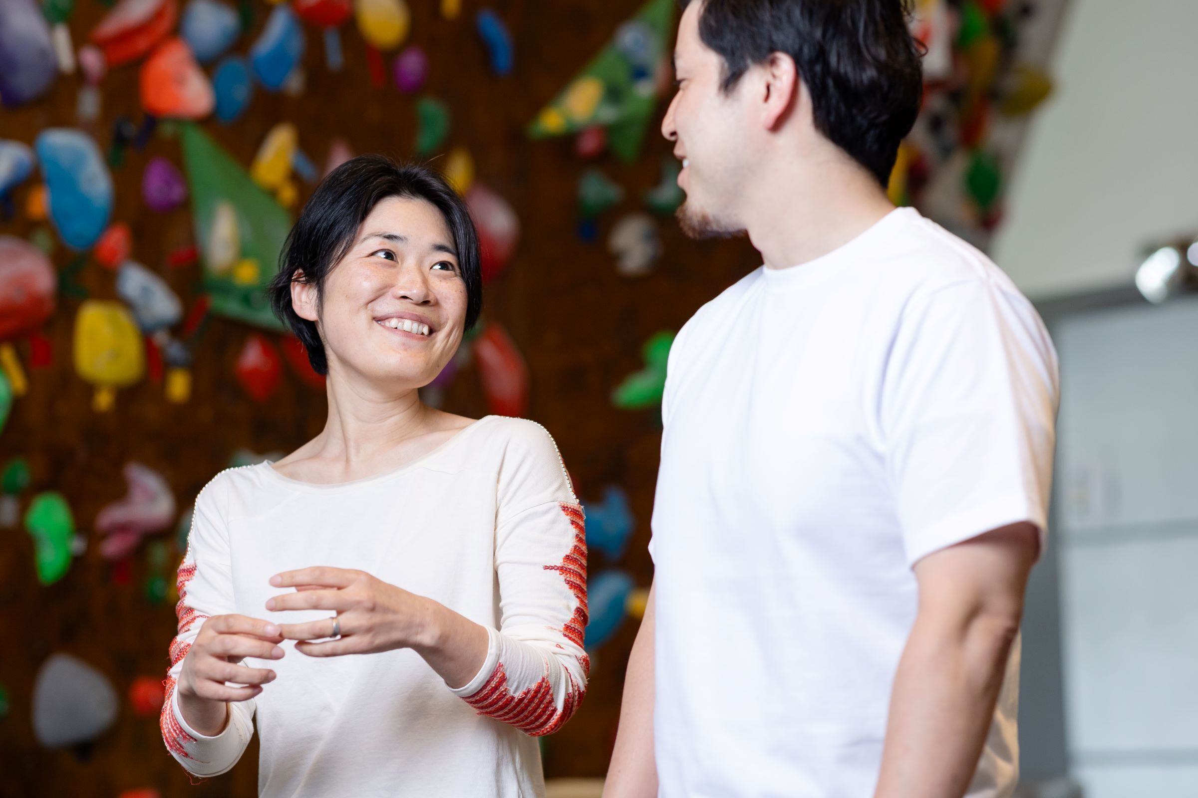 短髪の女性が微笑みながら隣に立っている白いTシャツを着ている男性を見ている。彼らの後ろにはカラフルなボルダリングウォールが見える。