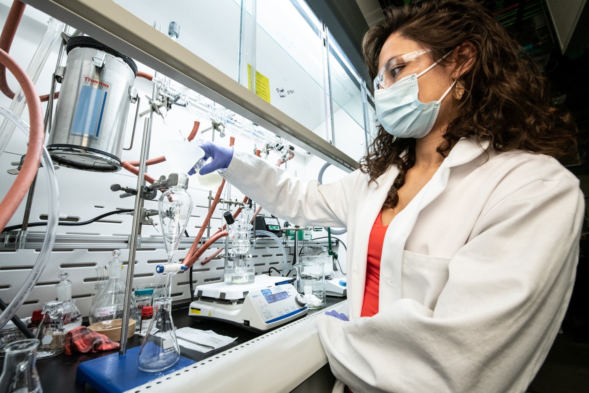 A woman scientist wears a mask and pours liquid into a glass beaker.