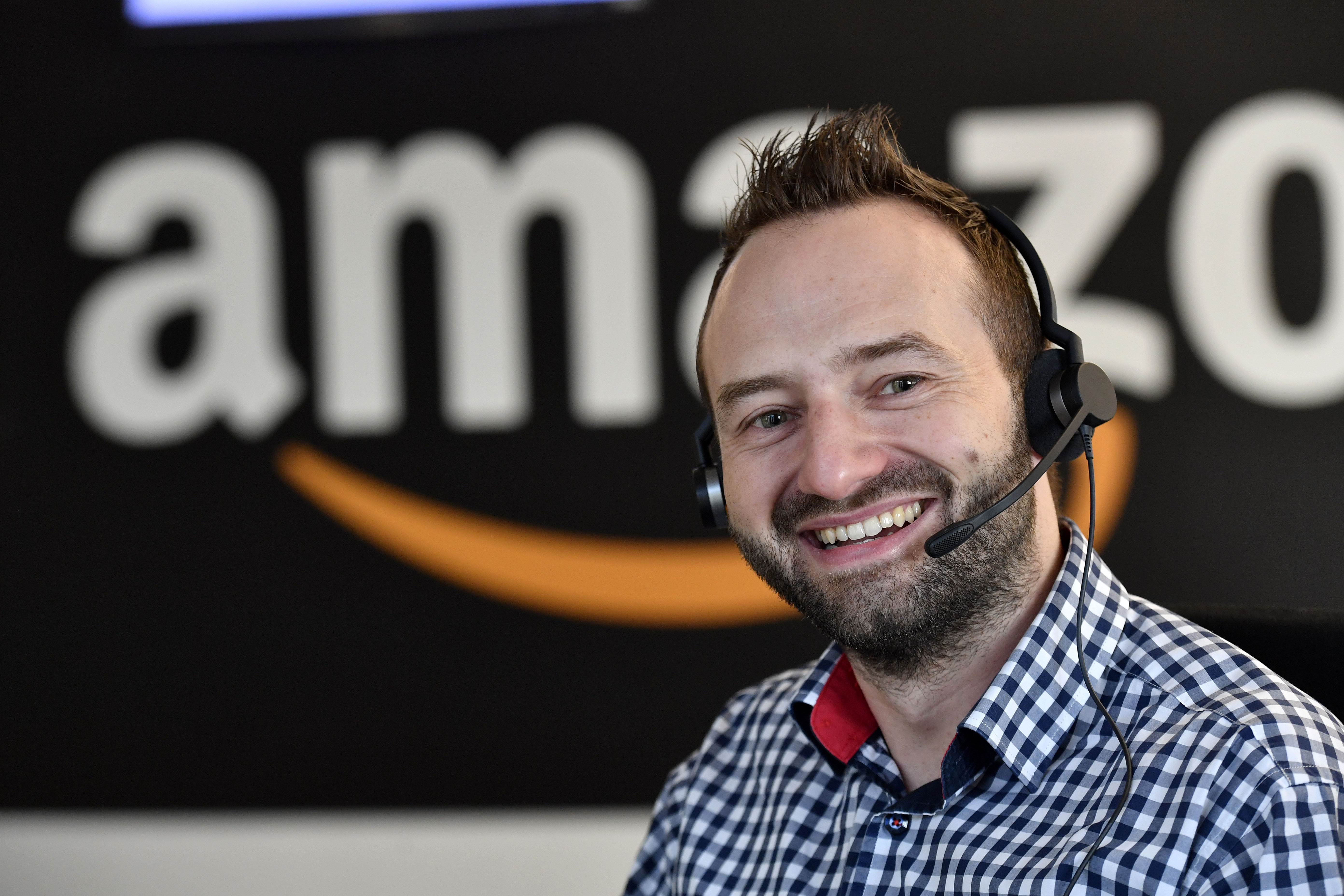 Ein dunkelhaariger Mann mit Bart und Headset lächelt in die Kamera. Er trägt ein kariertes Hemd. Im Hintergrund erkennt man etwas verschwommen das Amazon Logo.