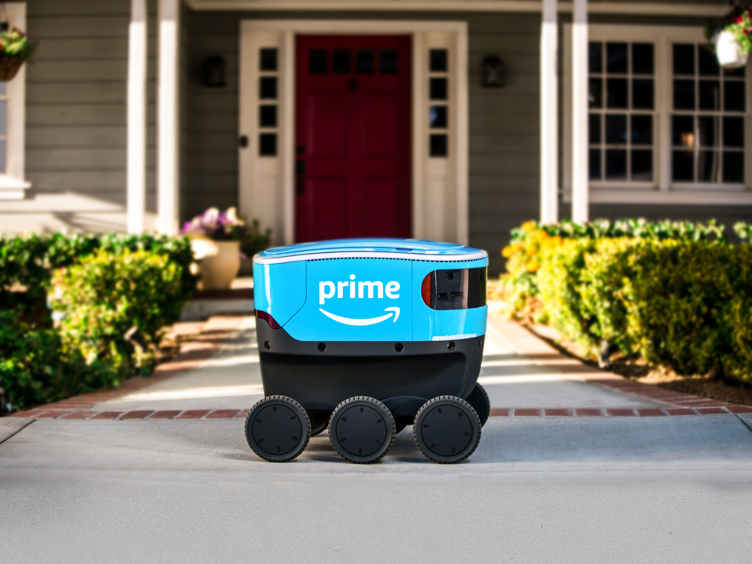 A bright blue, sleek, six-wheeled Amazon Scout delivery robot parked on the concrete walkway of an American home 