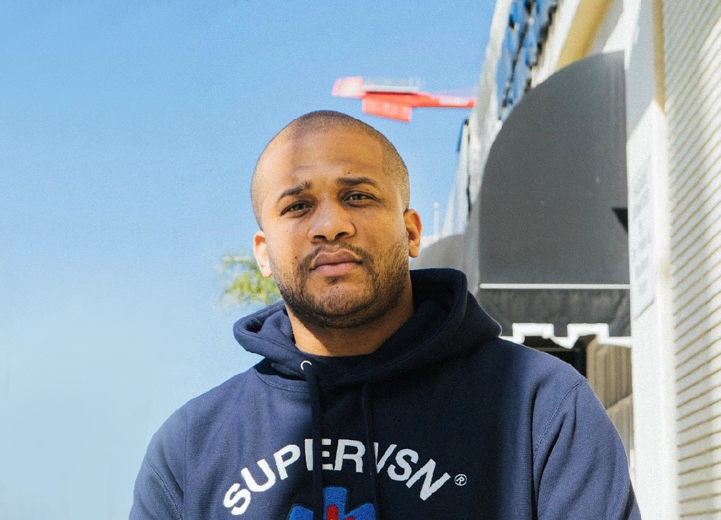 An image of a man standing for a photo outside. There is a palm tree and a street view behind him.