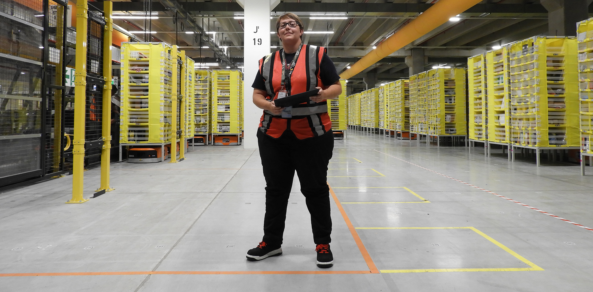 Die Amnesty Trainerin (schwarze Hose, grau-weiße Warnweste, Sicherheitsschuhe) ist in einer Logistikhalle zu sehen. Inder Hand hält sie ein Tablet. Hinter ihr sind zahlreiche gelbe Regale zu sehen, unter denen man Amazon Transportroboter erkennt.