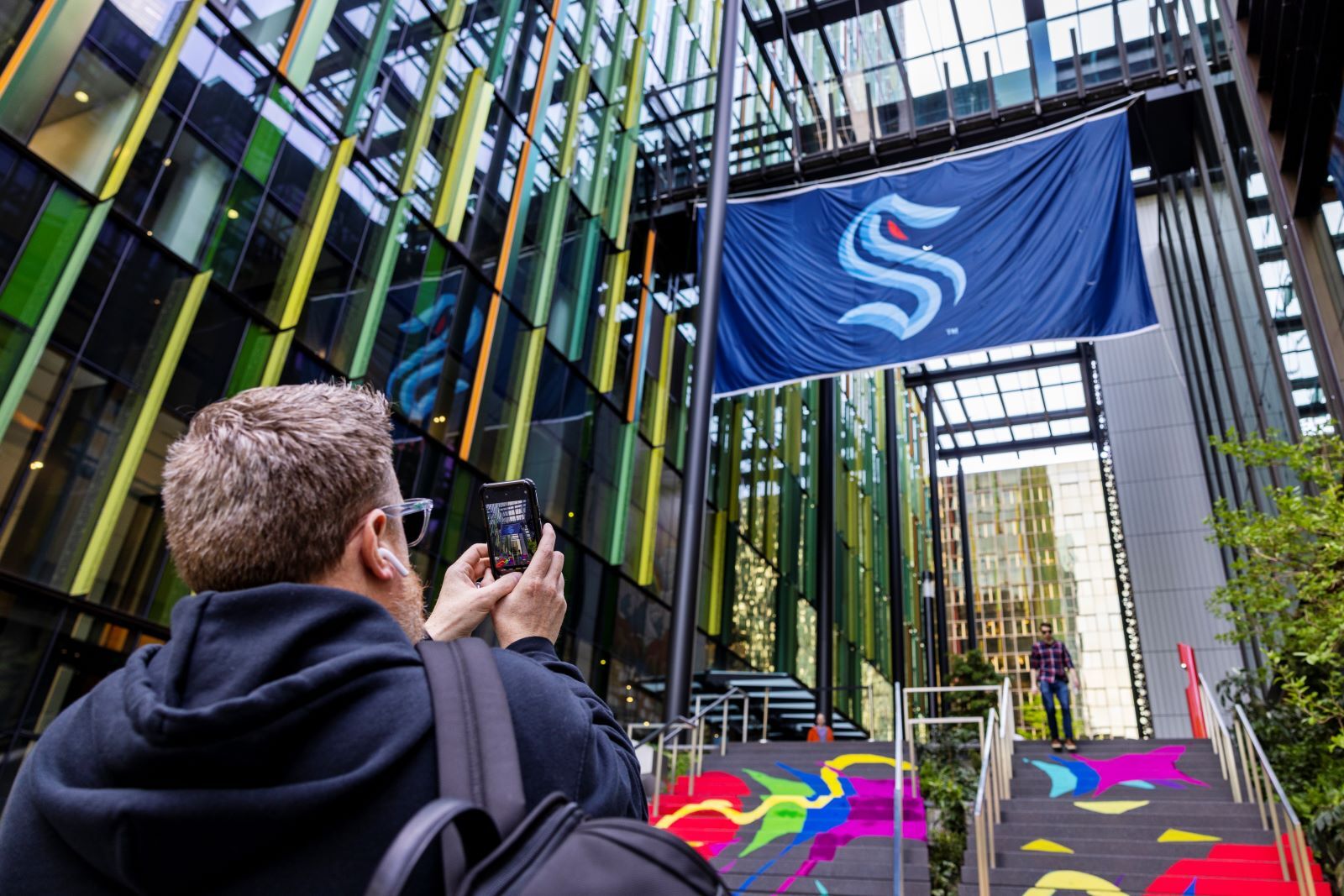 Man holding his phone up, taking a photo of a large Seattle Kraken flag that hangs between two buildings in downtown Seattle.