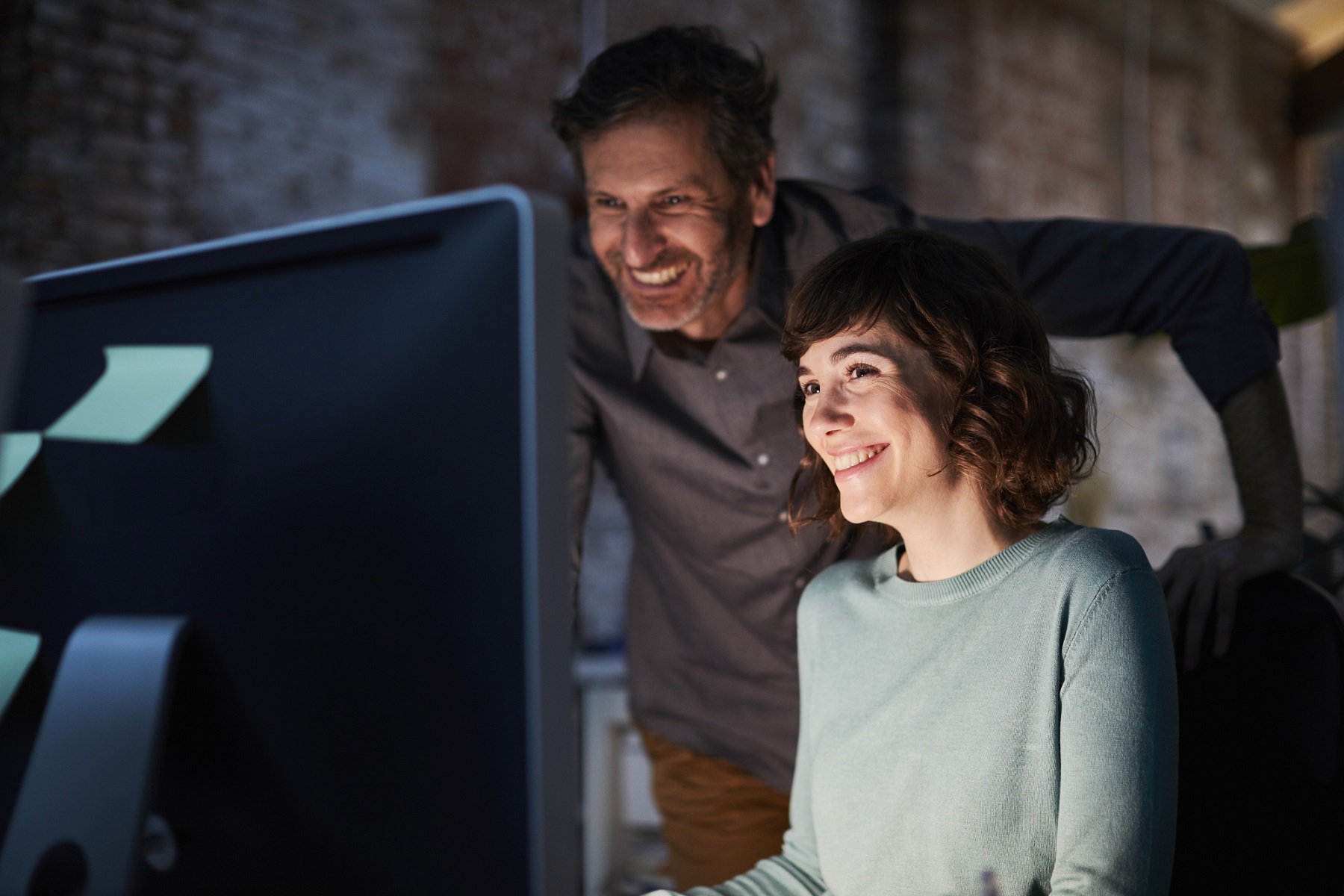 Man and woman looking at a computer screen. Both individuals are smiling. The light from the monitor lights their faces in an otherwise dark room. 