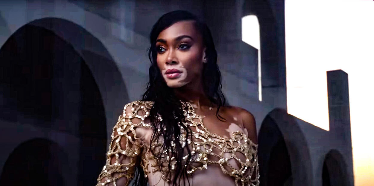 An image of model Winnie Harlow standing in a breezeway of a stone building. She is looking off into the distance.
