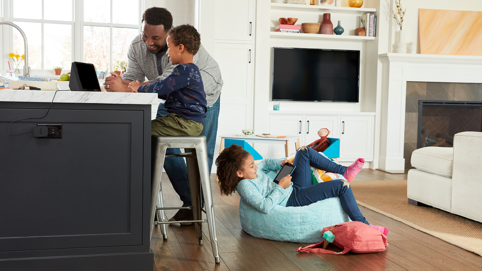 An image of a family interacting with Amazon devices like an Echo Show and a Kindle in their living room. The dad and son are sitting on the kitchen stools and the daughter is sitting on a light blue beanbag with a pink backpack next to her. 