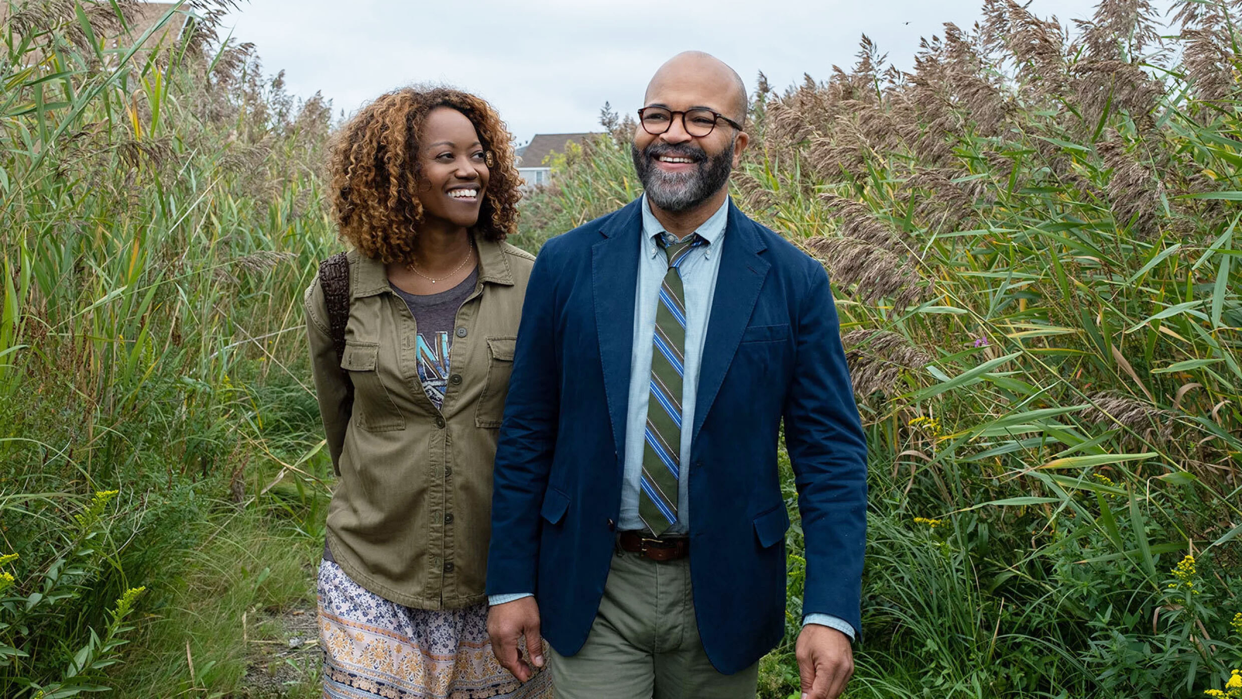 Actors Jeffrey Wright and Erika Alexander walking through tall green bushes, smiling, and holding hands in a scene from the movie, American Fiction. 