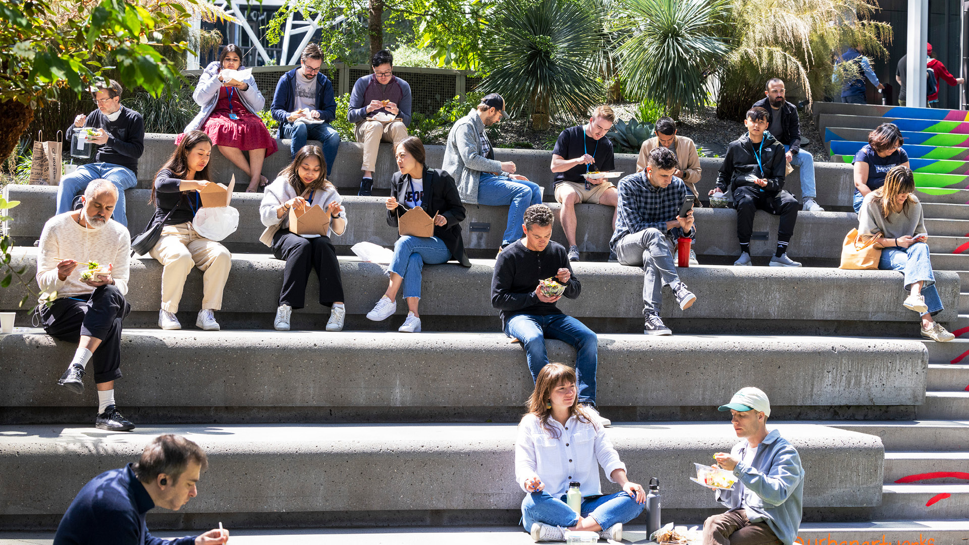 Several Amazon employees sitting on the concrete stairs outside of an Amazon building in Seattle. Many of them are seated in groups, eating lunch from takeout containers.