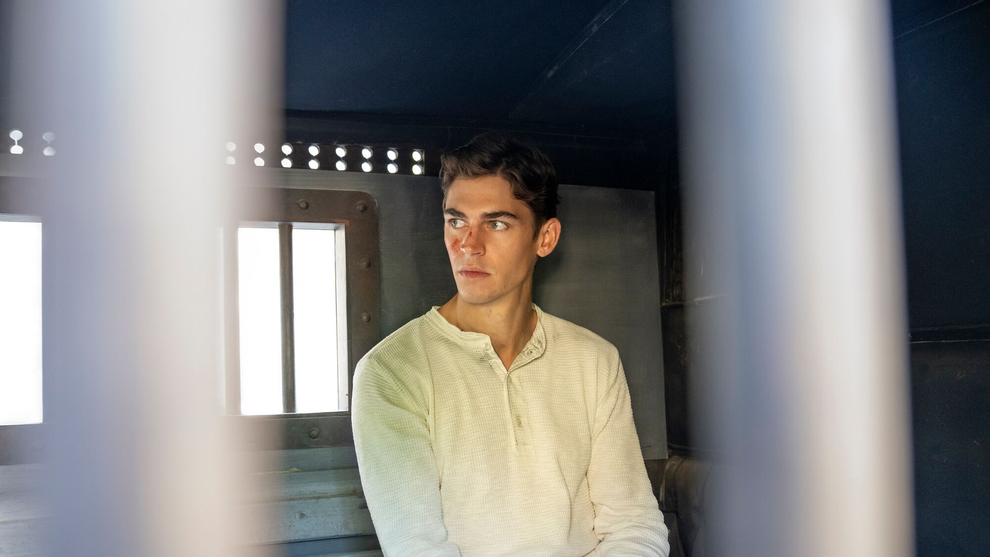 Hero Fiennes Tiffin as Young Sherlock in white henley shirt gazing off-camera in dimly lit box.