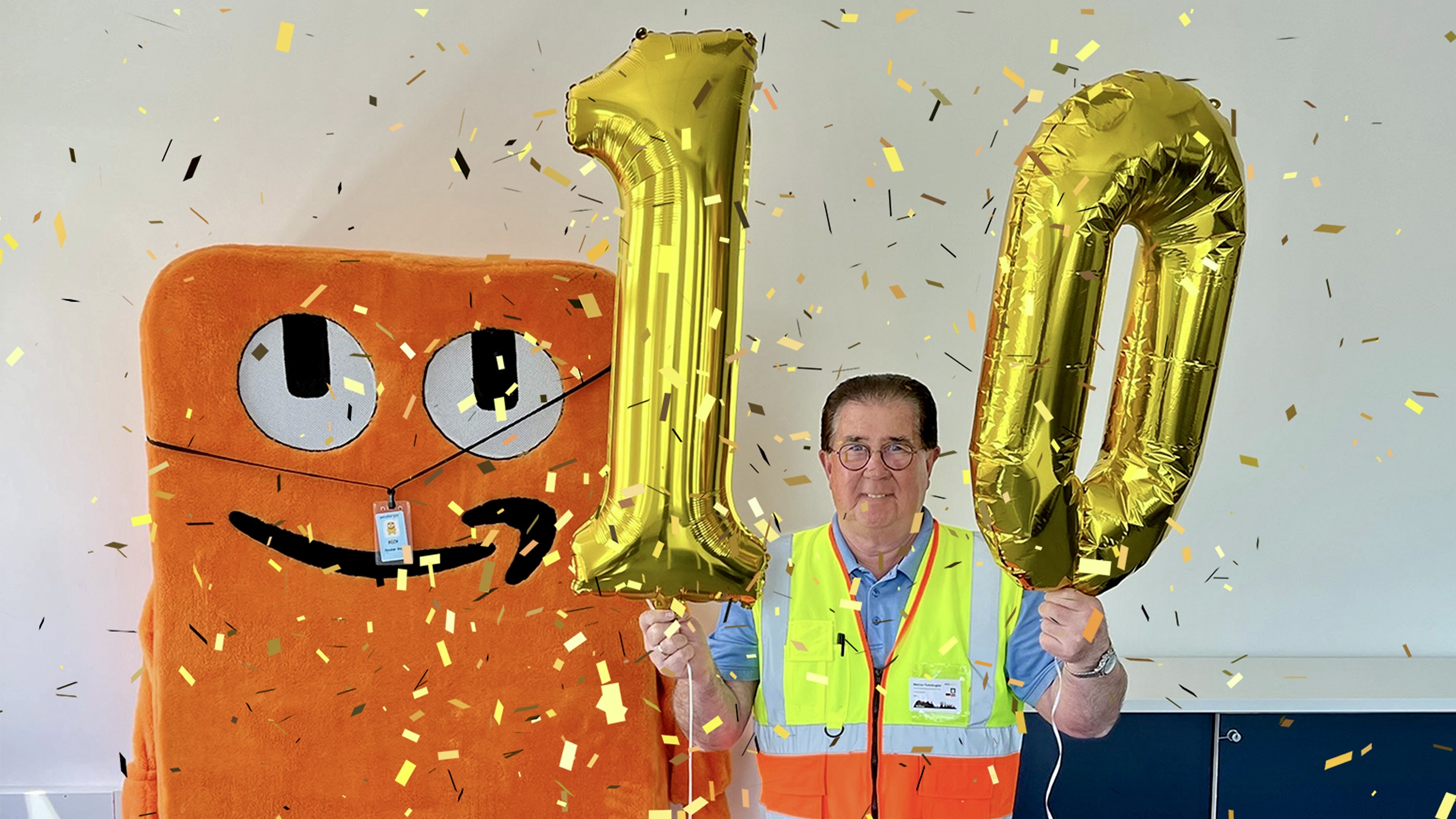 Amazon Logistikmitarbeiter Werner Ramskugler neben dem Amazon Maskottchen Peccy: Er hält Luftballons mit einer 1 und einer 10 in der Hand.