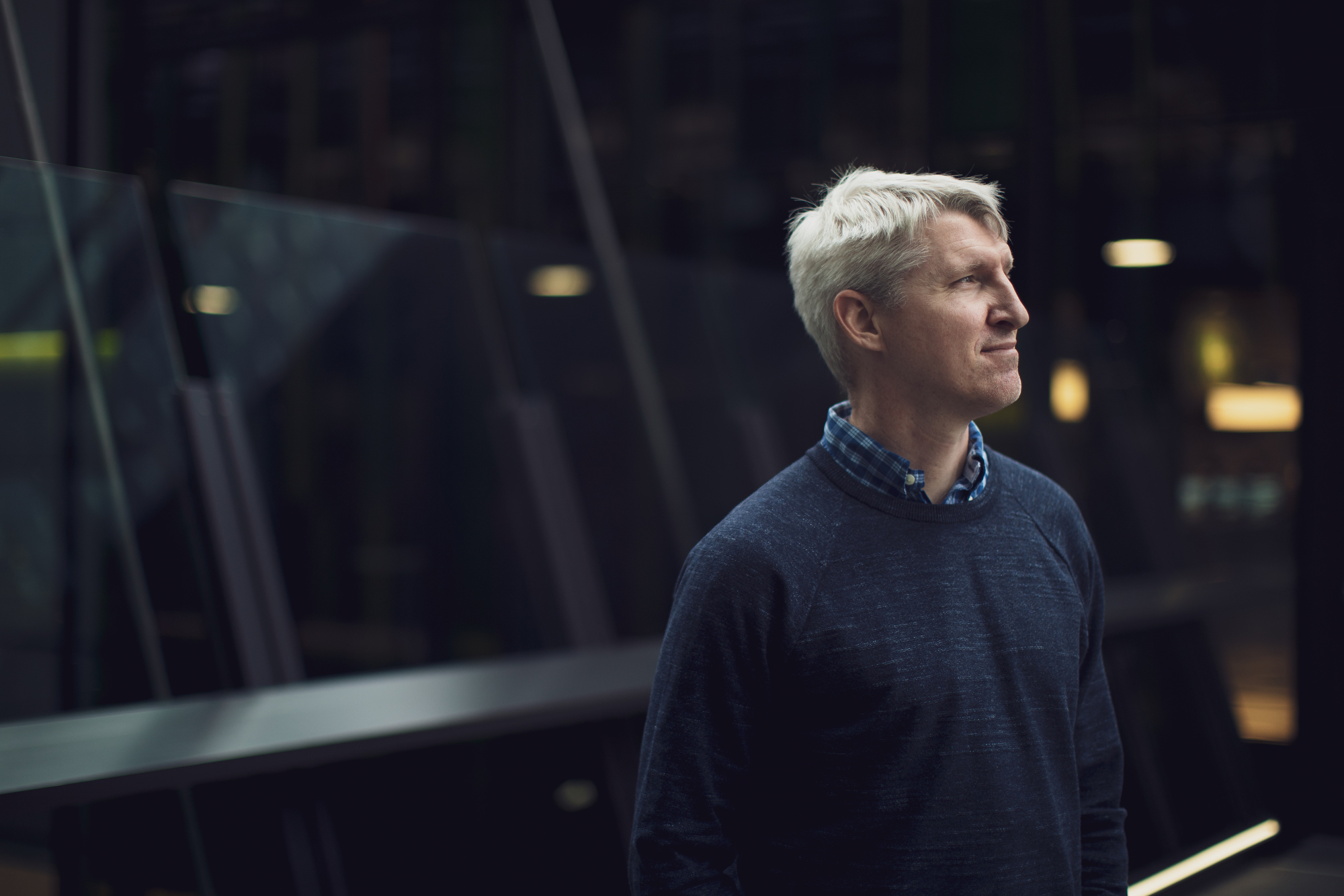 A man stands in a covered walkway near a wall of glass. He looks away from the photographer, as the photo catches his profile.