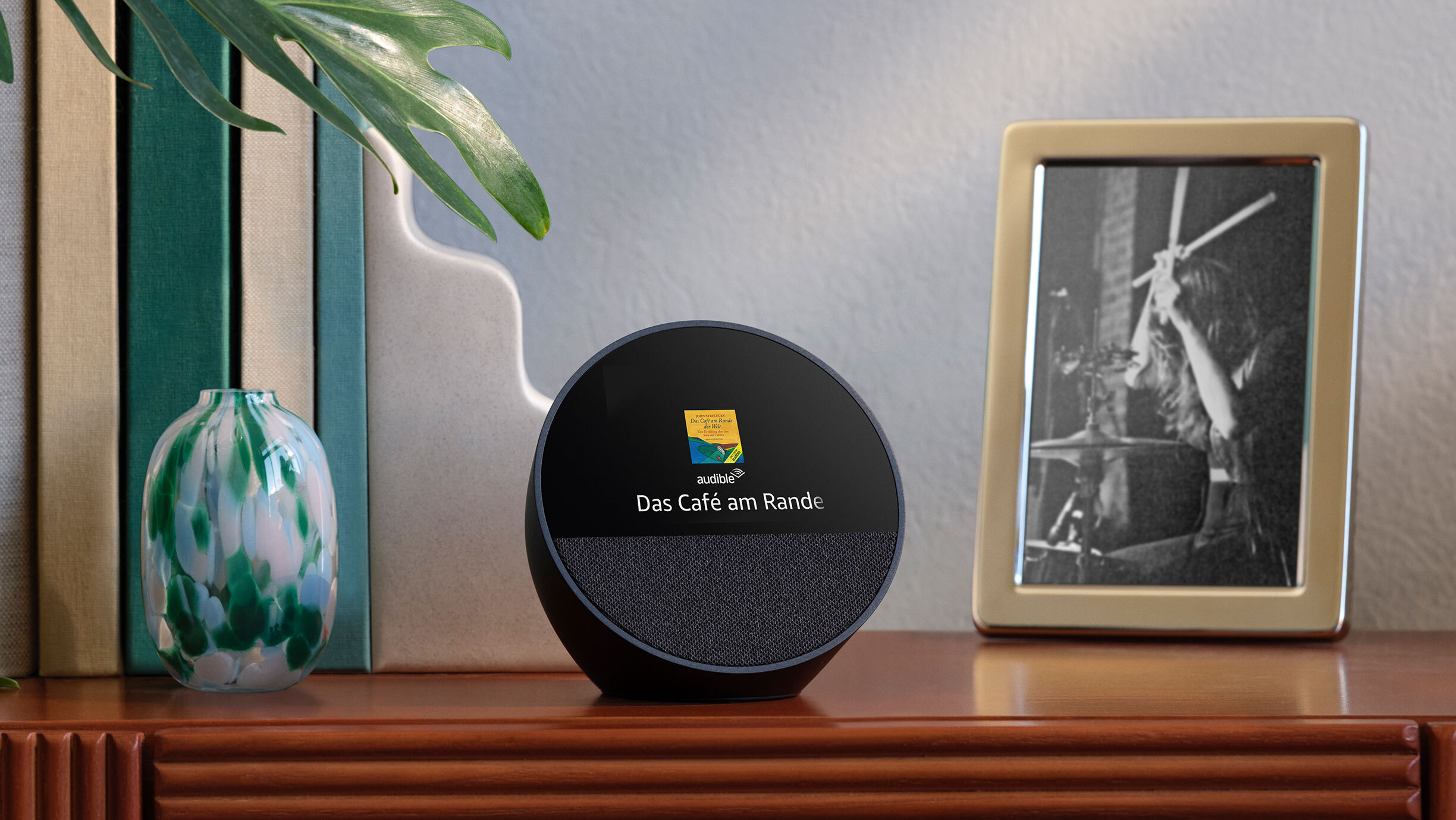 Ein schwarzer Echo Spot auf einem Siteboard, links steht eine bunte Vase, rechts ist ein Schwarzweiß-Bild mit goldenem Rahmen zu sehen. Der Echo zeigt einen Teil des Buchtitels: "Das Café am Rande ..." 
