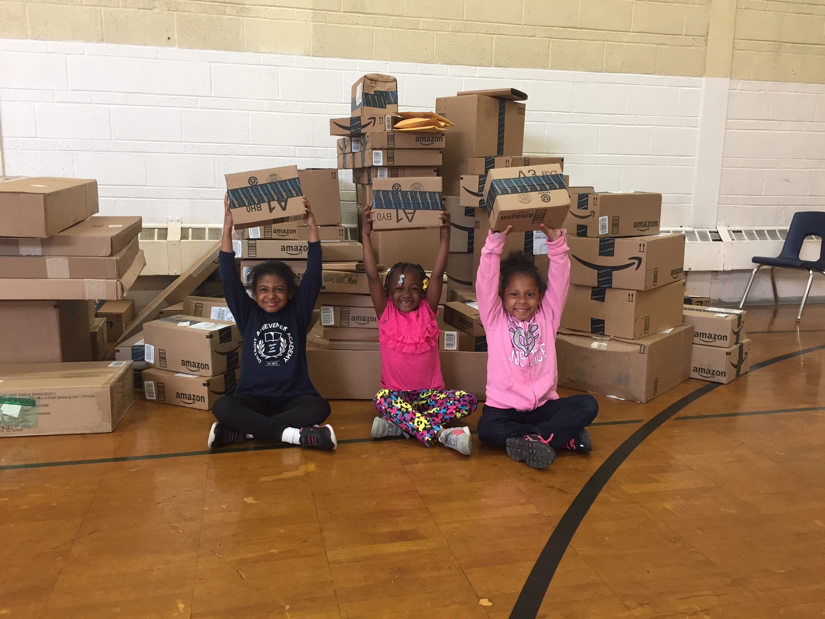 Three children holding Amazon delivery boxes over their hands and smiling. They are in a school gymnasium in Detroit, surrounded by Amazon boxes.