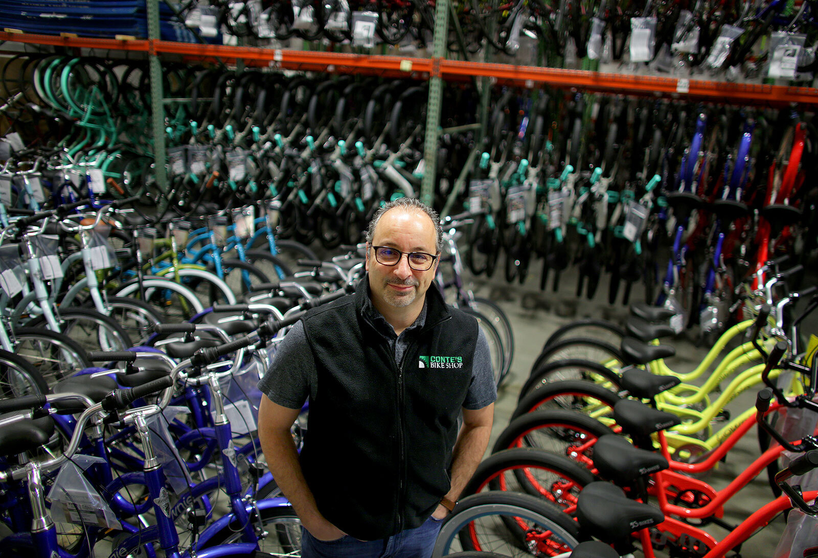 An image of a man smiling for a photo in a bike shop with rows of bikes behind him. He is wearing a vest that says "Conte's Bike Shop."