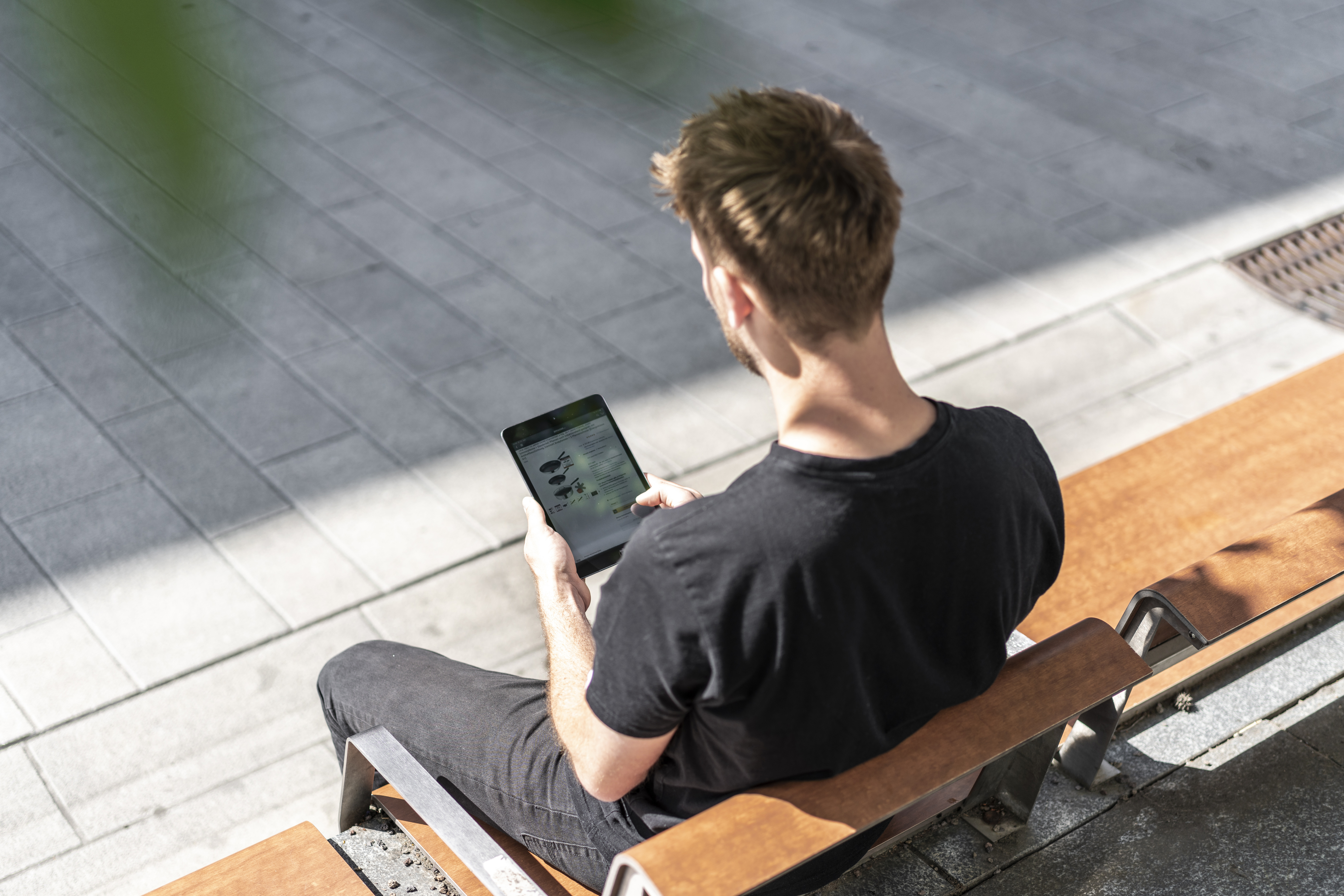 Ein junger Mann sitzt auf der Parkbank und hat ein Tablet in der Hand. Er bestellt von unterwegs bei Amazon.