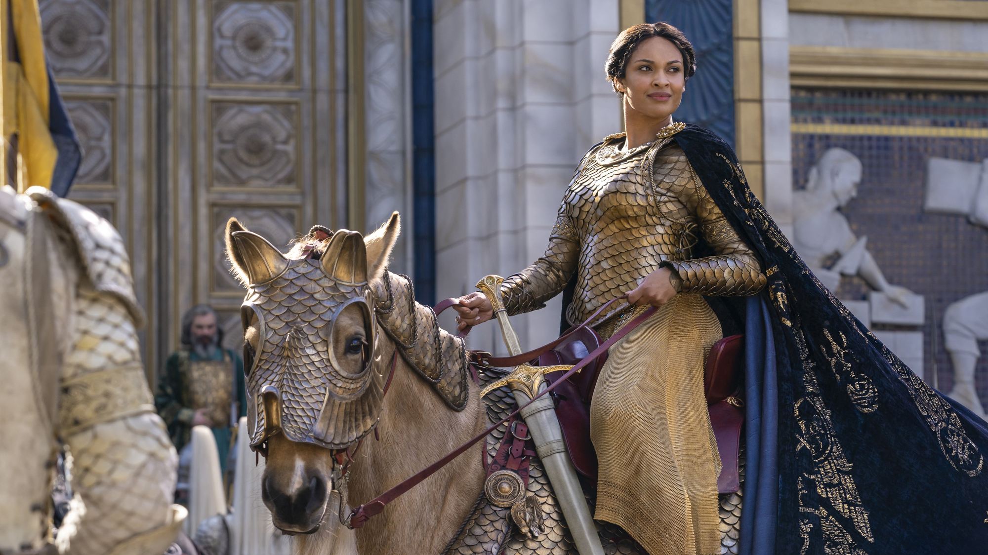 A woman riding on a horse in front of a marble building wears an outfit of gold armor.