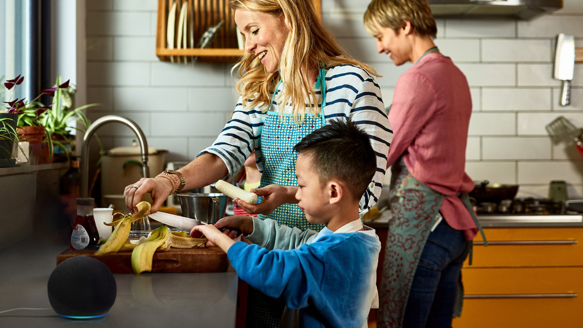 An image of two adults and a child cooking in a kitchen and peeling bananas. There is an Amazon Echo device sitting on the kitchen counter. 