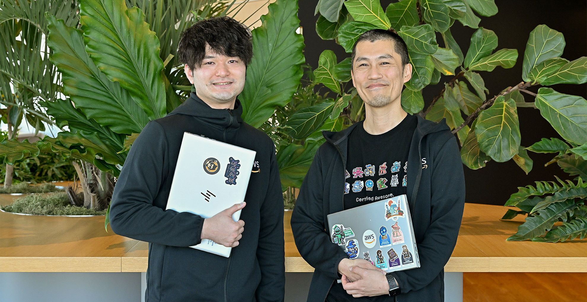 黒いシャツを着ている2人の男性がパソコンを持ちながら笑っている