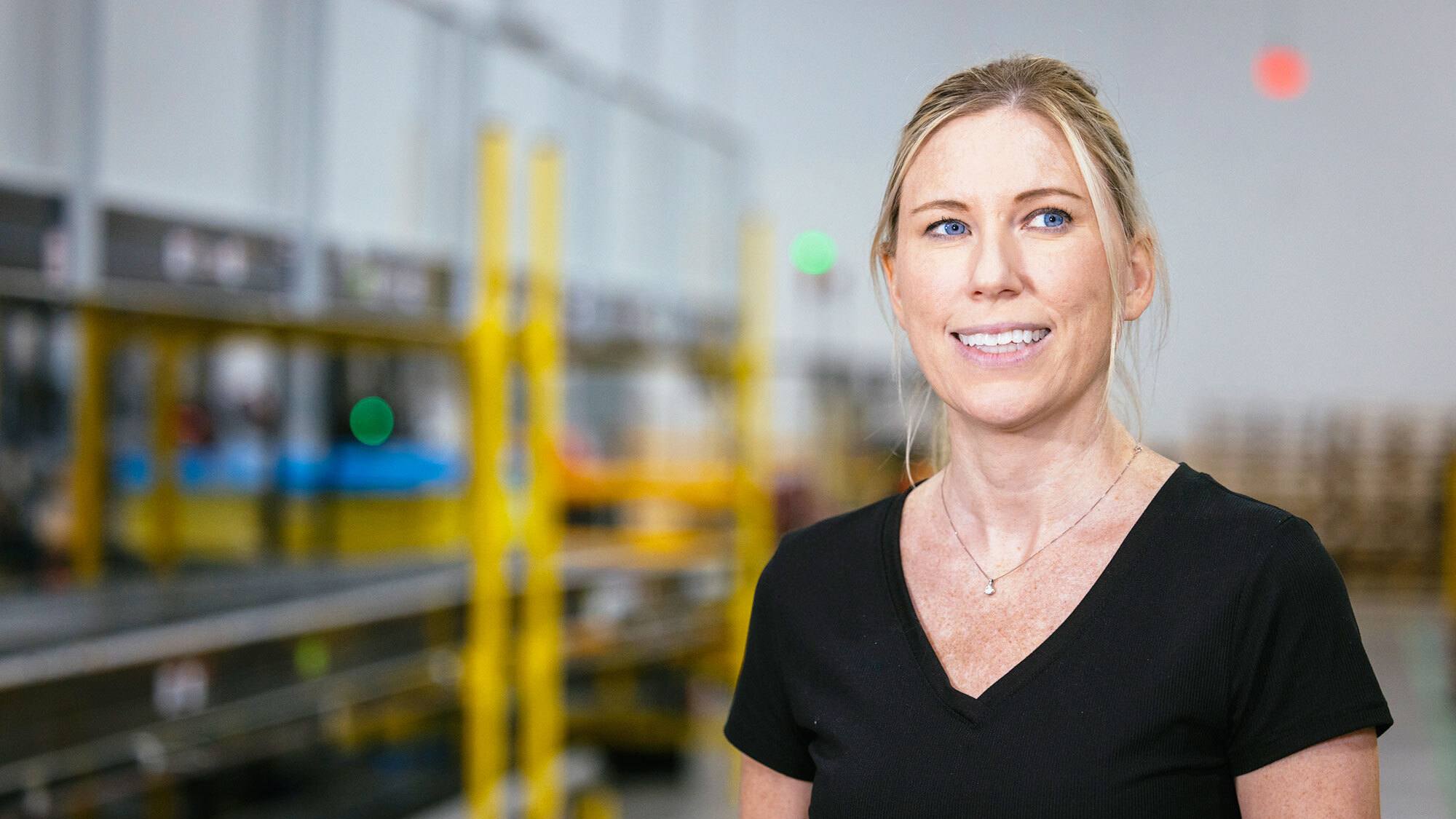A woman wears a black T-Shirt and smiles as she poses for a picture in an Amazon fulfillment center.