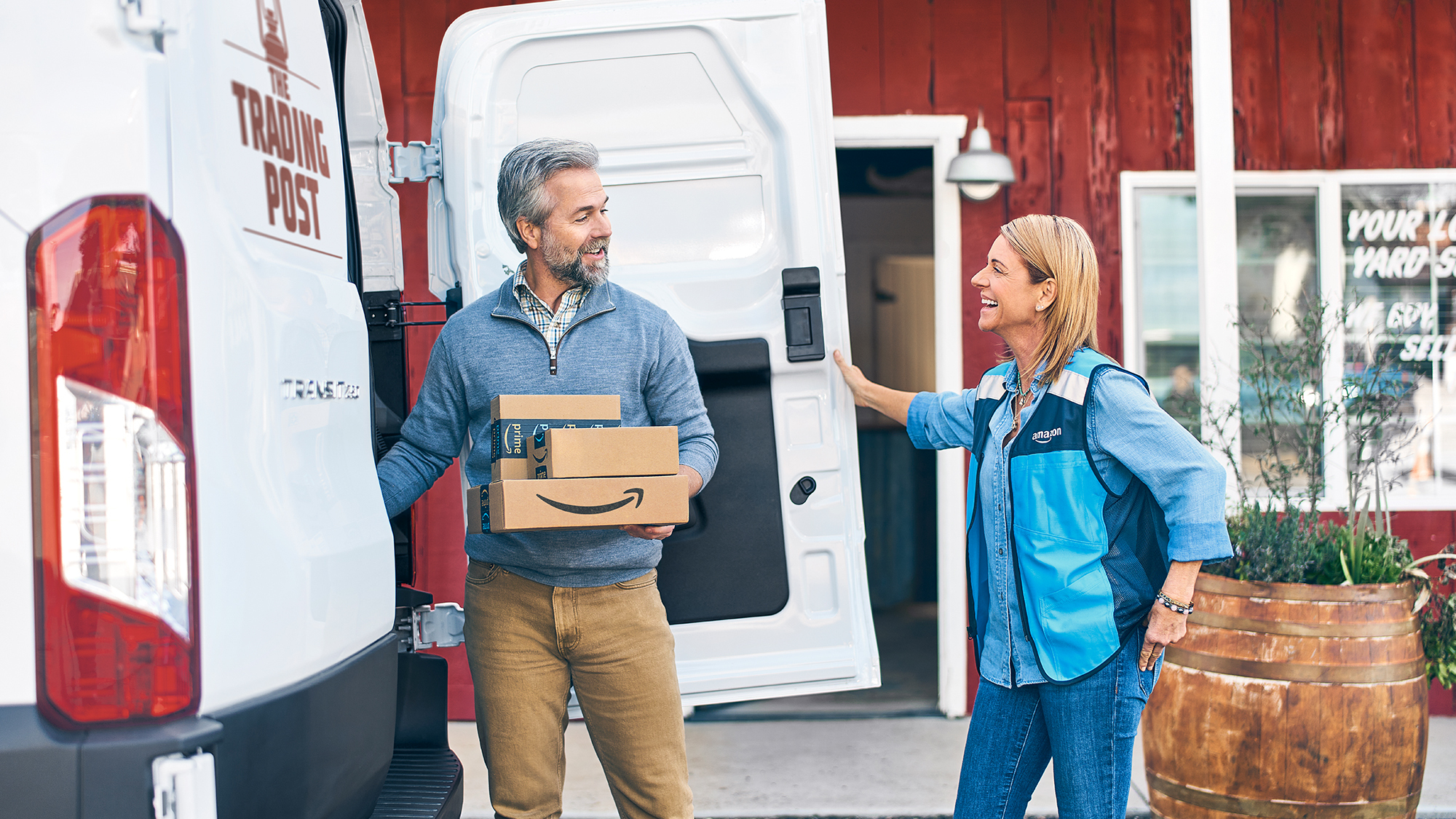 An Amazon employee wears a blue Amazon vest and talks with a customer while he unloads packages from a work van at his store.