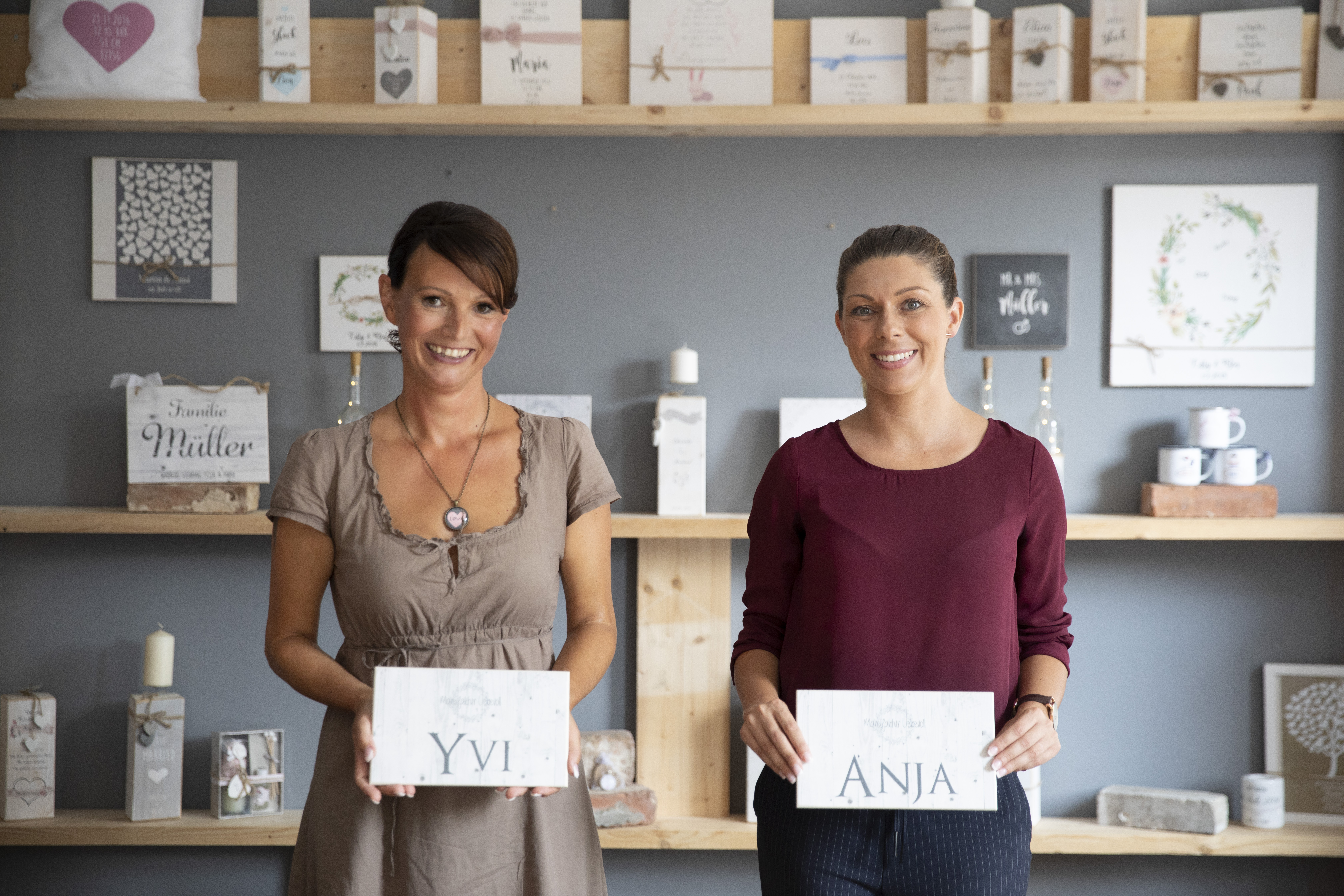 Zwei Frauen mit dunklen Haaren stehen vor einer grauen Wand und schauen in die Kamera. In ihren Händen halten sie weiße Schilder mit ihren Namen "Yvi" und "Anna" in die Luft.