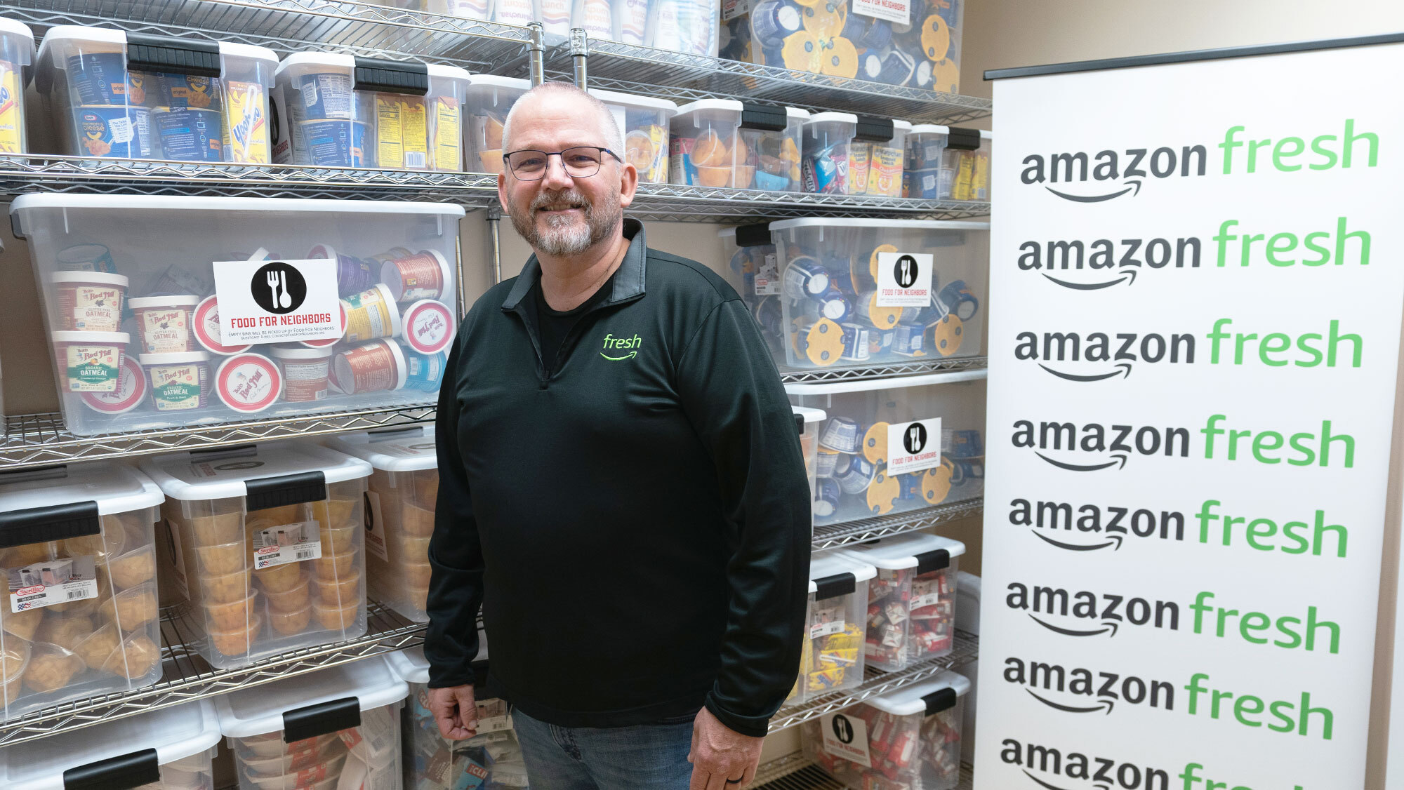 An image of a person standing in front of an Amazon Fresh banner with packages of food in the background. 