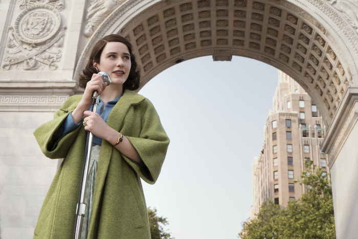 An image of Mrs. Maisel standing in front of the Washington Square Arch in New York. She is wearing a green coat and a blue top. There is a microphone in front of her and she is speaking to the crowd. 