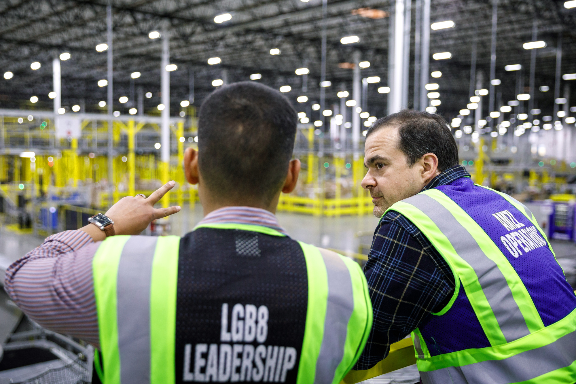 A man wearing a safety vest is explaining something to another man on his right (Jeff Wilke, CEO of worldwide consumer at Amazon), both men are wearing safety vests. In the background, an Amazon fulfillment center. 