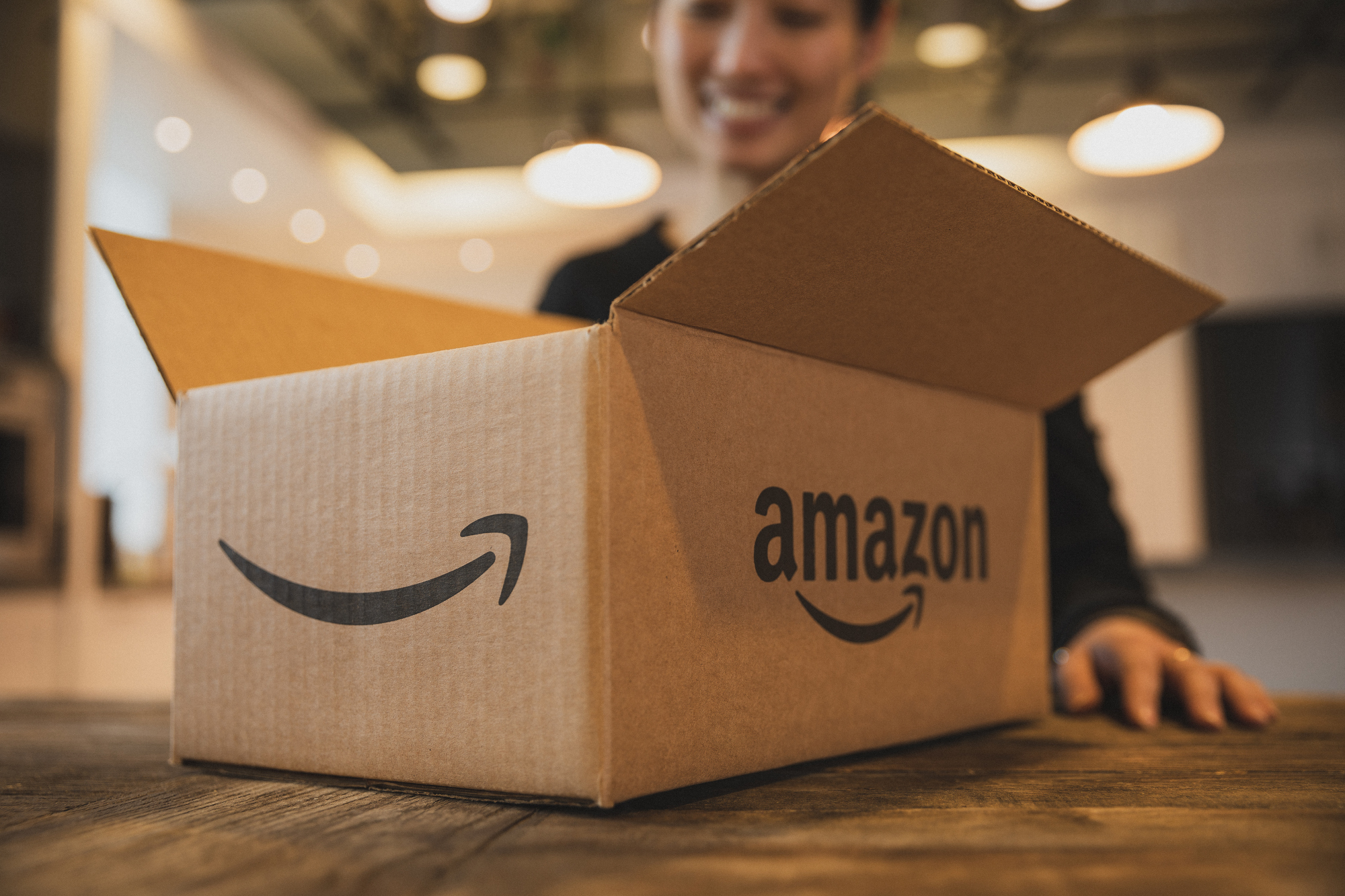 Geöffnetes Amazon Packet auf einem Tisch, wobei im Hintergrund eine junge Frau zu sehen ist.