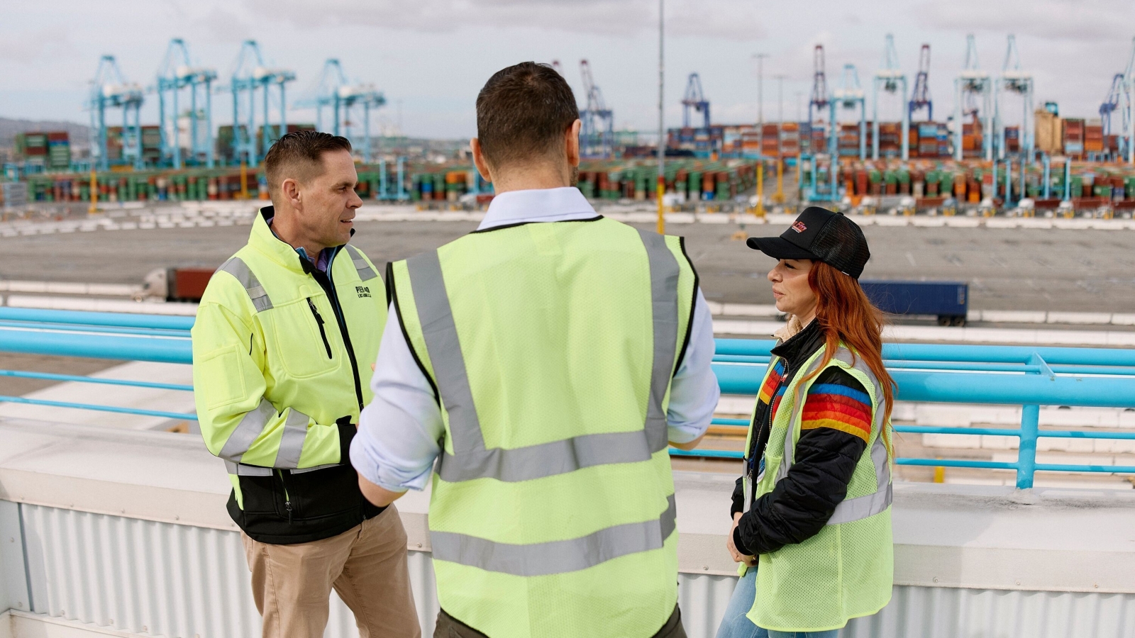 Three Amazon employees in safety vests discussing operations at a busy shipping container port