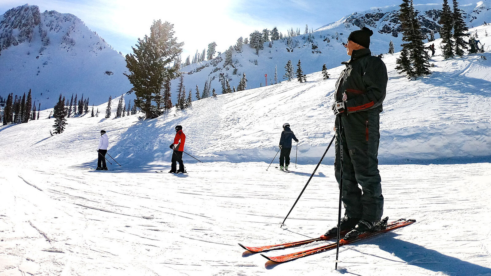 An image of a man on a ski slope. He is wearing a dark grey ski suit with his skis strapped on and his poles in either hand. He is smiling and looking off towards the bright blue sky with sun shining down.