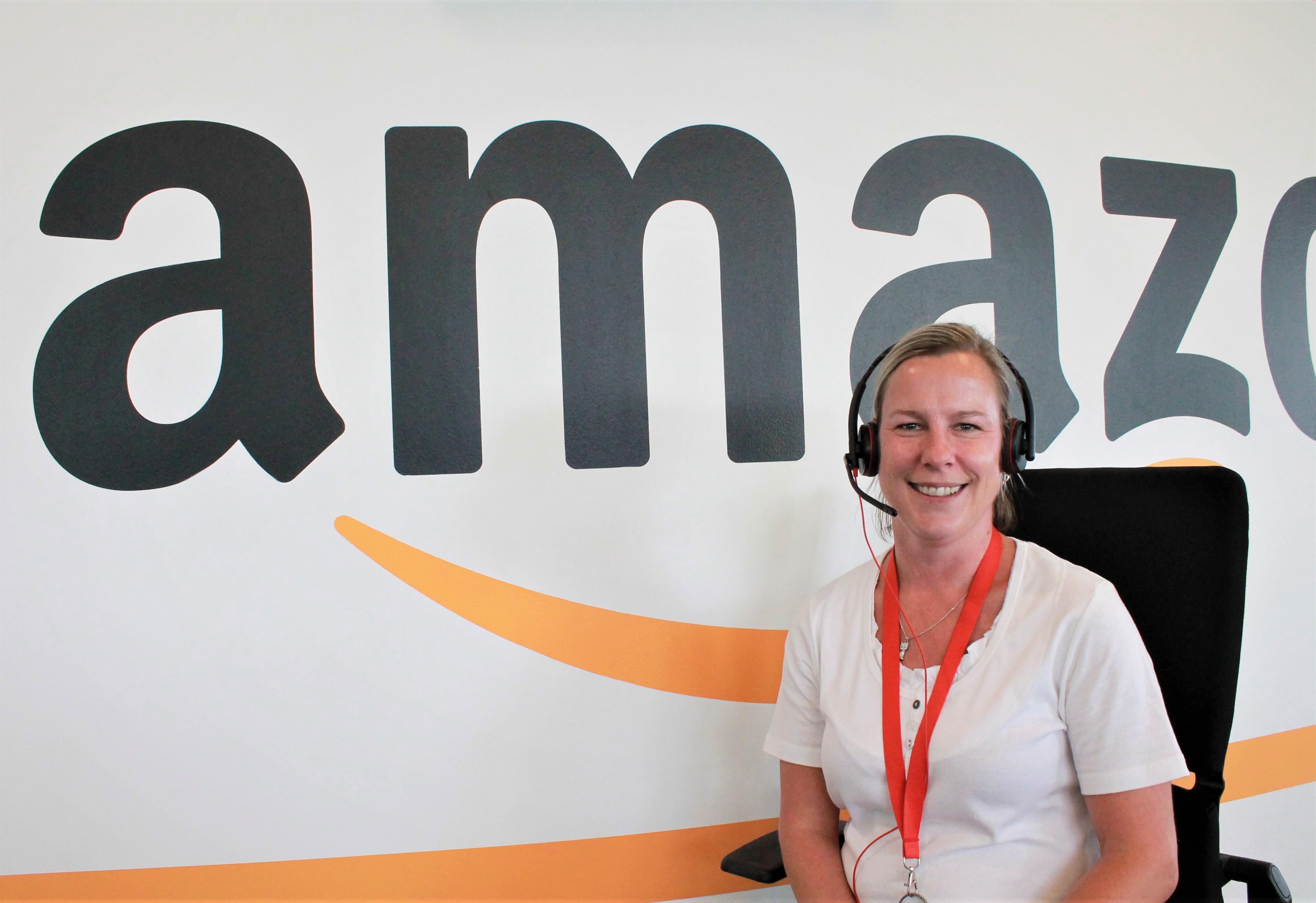 Eine Amazon Kundenservice-Mitarbeiterin mit Kopfhörer vor einer Wand mit Amazon Schriftzug.