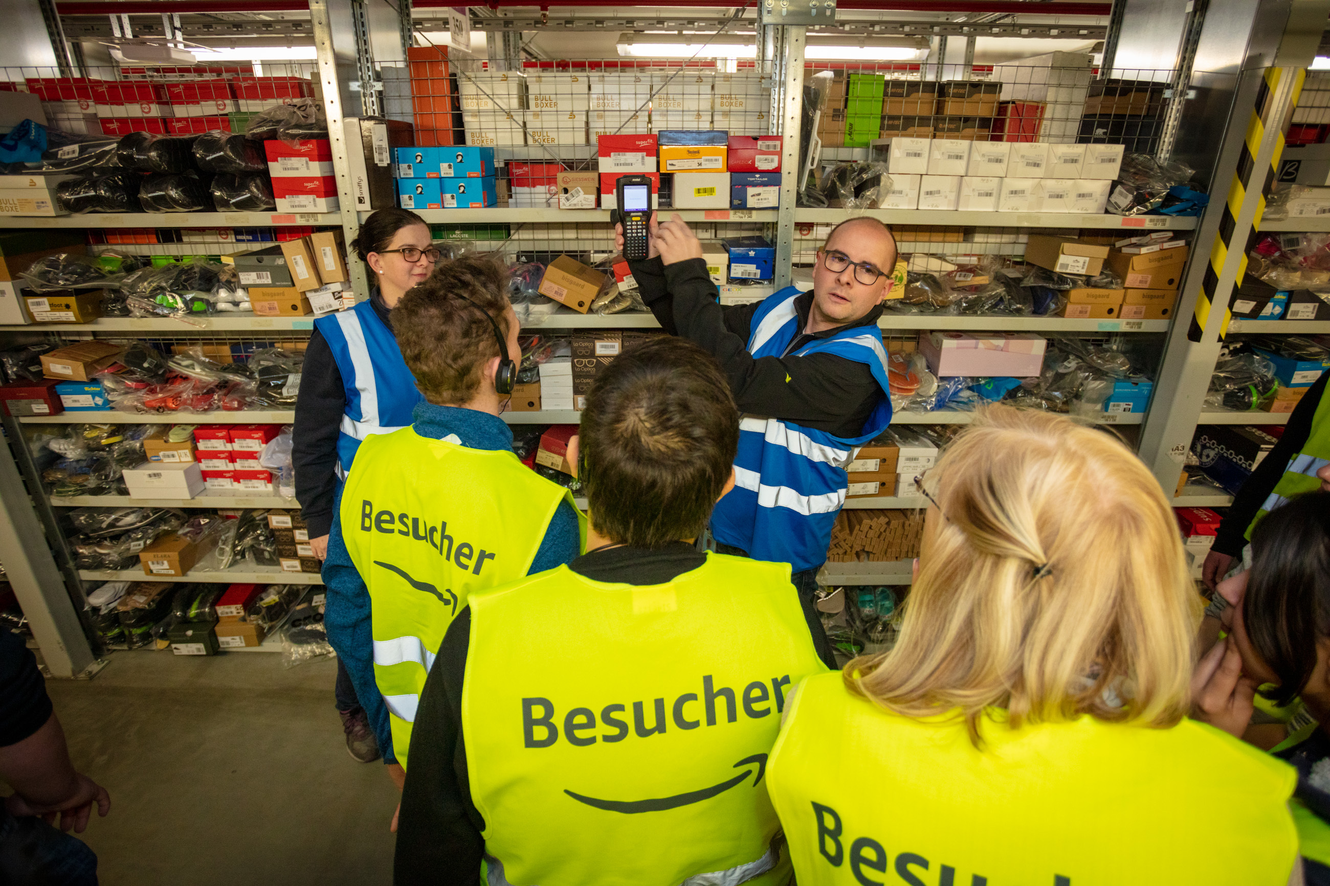 3 jugendliche Besucher (von hinten sichtbar) stehen vor 2 Amazon Mitarbeiter, die die Scanner-Funktion erklären.