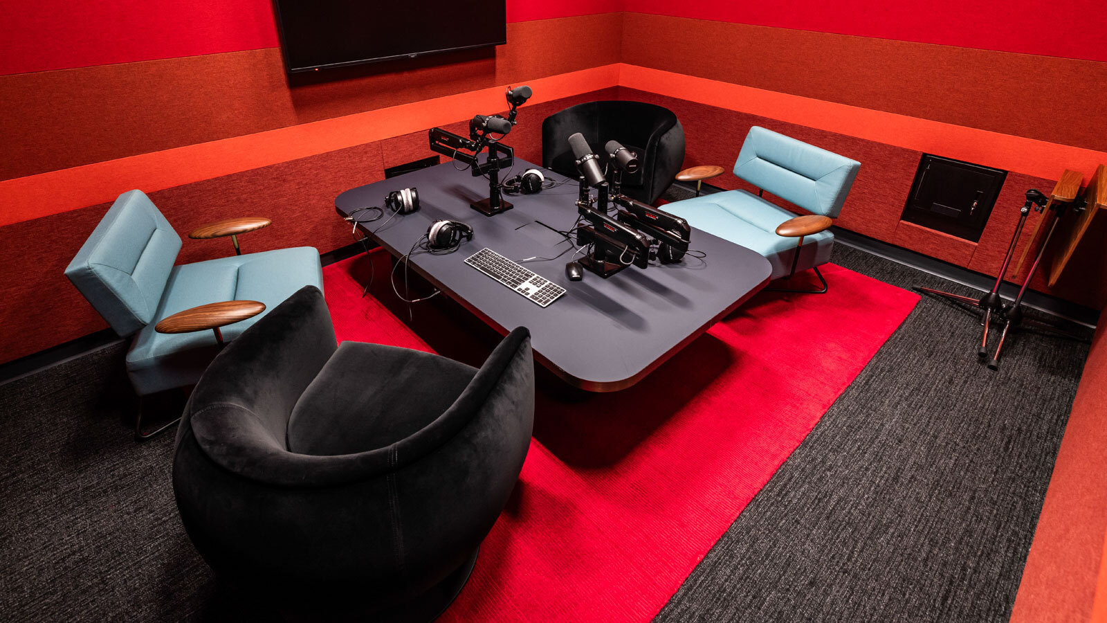 A red and grey room set up for podcast recording. There are four large, comfy chairs pulled up to a table with microphones and a TV on the wall.