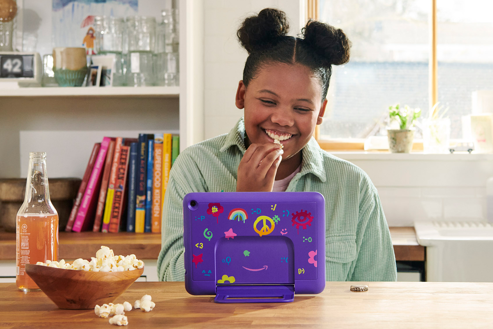 A young girl smiles as she watches her Fire Kids tablet that is protected with a purple case decorated with stickers. She is eating popcorn and has an orange soda.