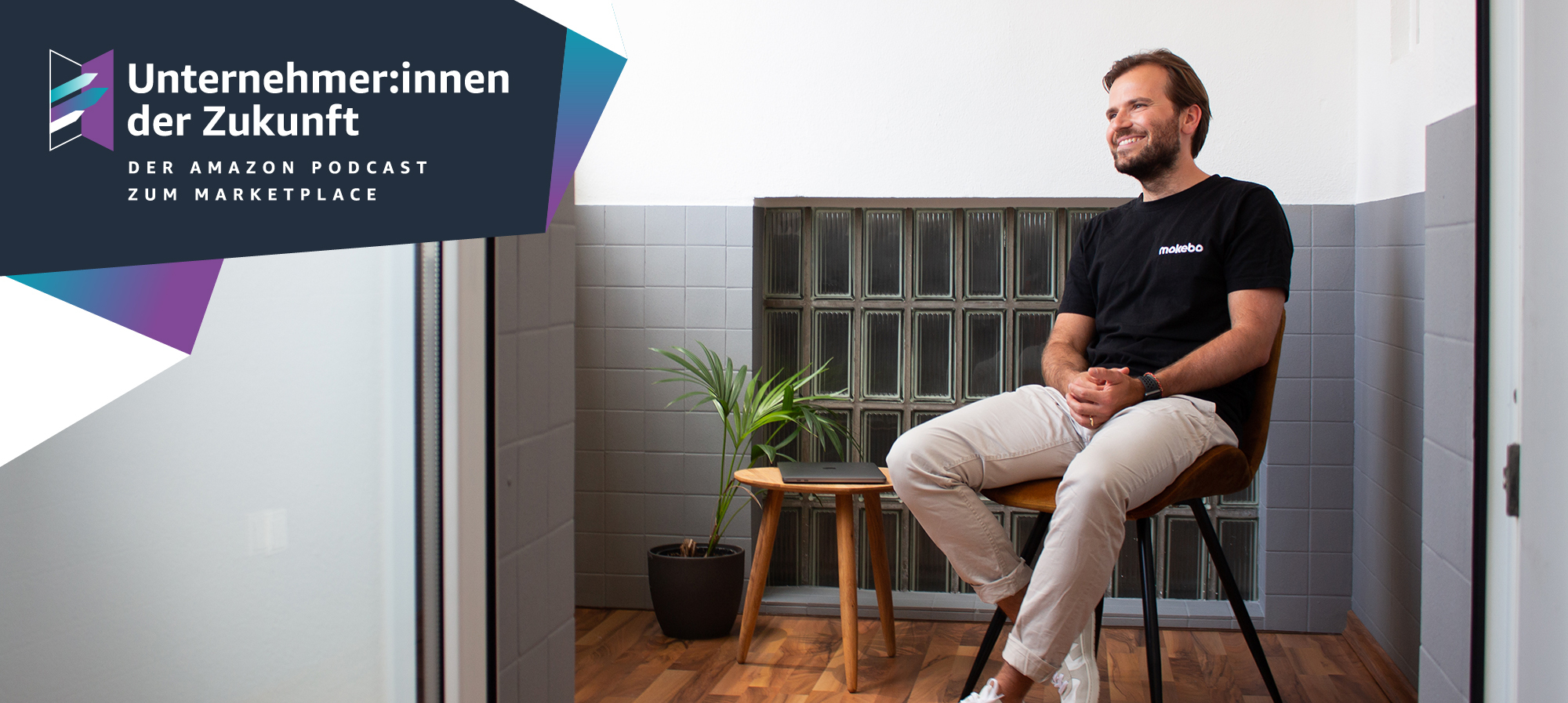 Ein Mann mit braunen Haaren, schwarzem T-shirt und heller Hose sitz auf einem Stuhl und lacht.