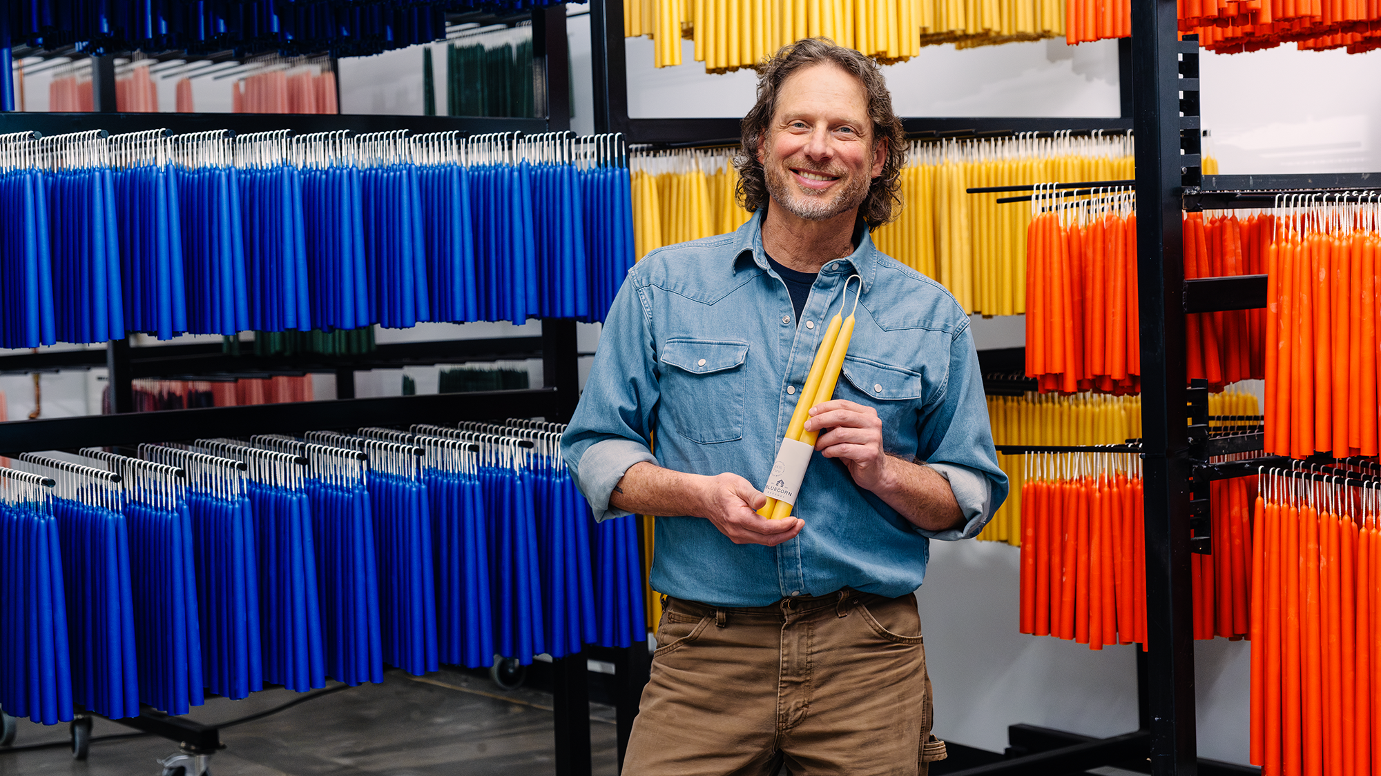 A man holding a pair of candles. There are hanging beeswax candles hanging behind him.