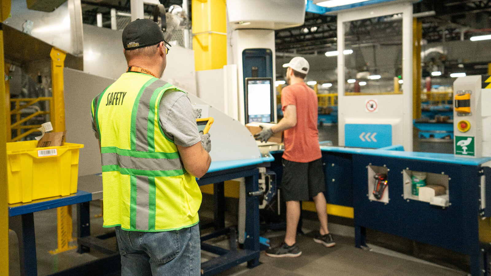 An image of an Amazon employee with a yellow safety vest on watching assisting another employee in safety practices while they work with a robotic system in an Amazon Fulfillment center. 