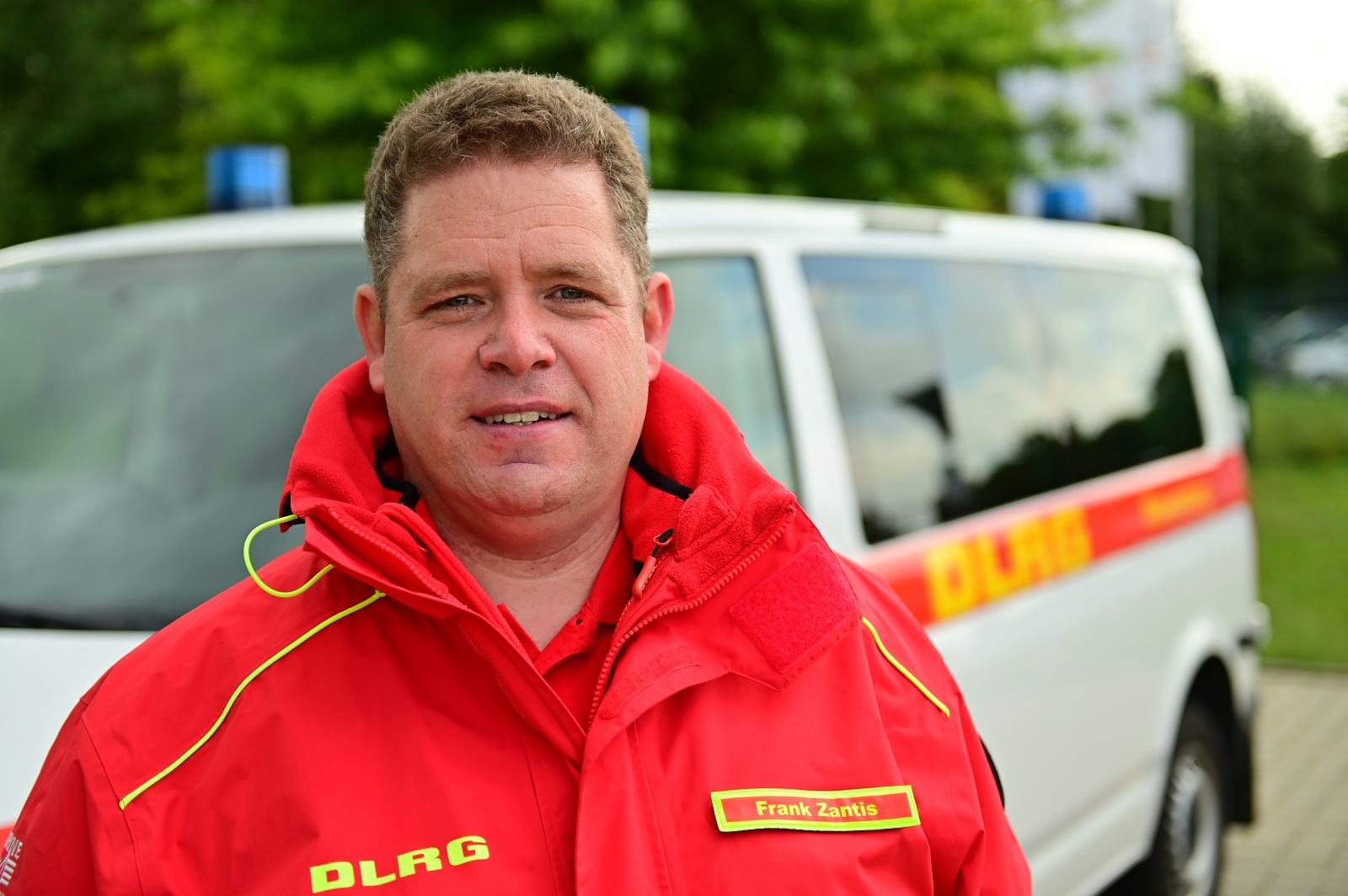 Frank Zantis steht vor einem Einsatzwagen der DLRG. Er trägt die rote Einsatzkleidung der Deutschen Lebensrettungsgesellschaft.