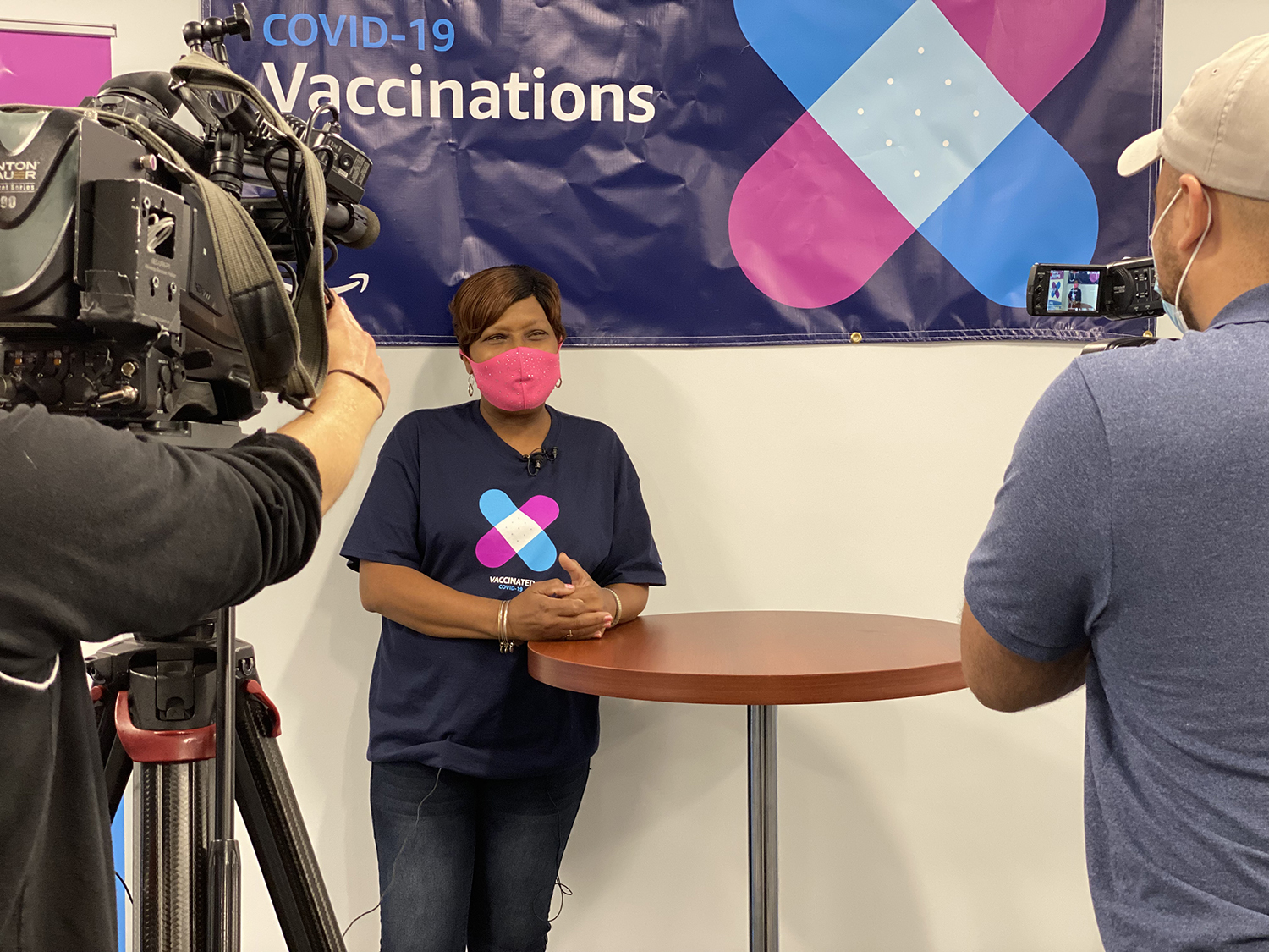 An image of a woman wearing a pink mask while smiling and leaning against a tall table. She is wearing a shirt showing she has been vaccinated and being filmed by a film crew. 