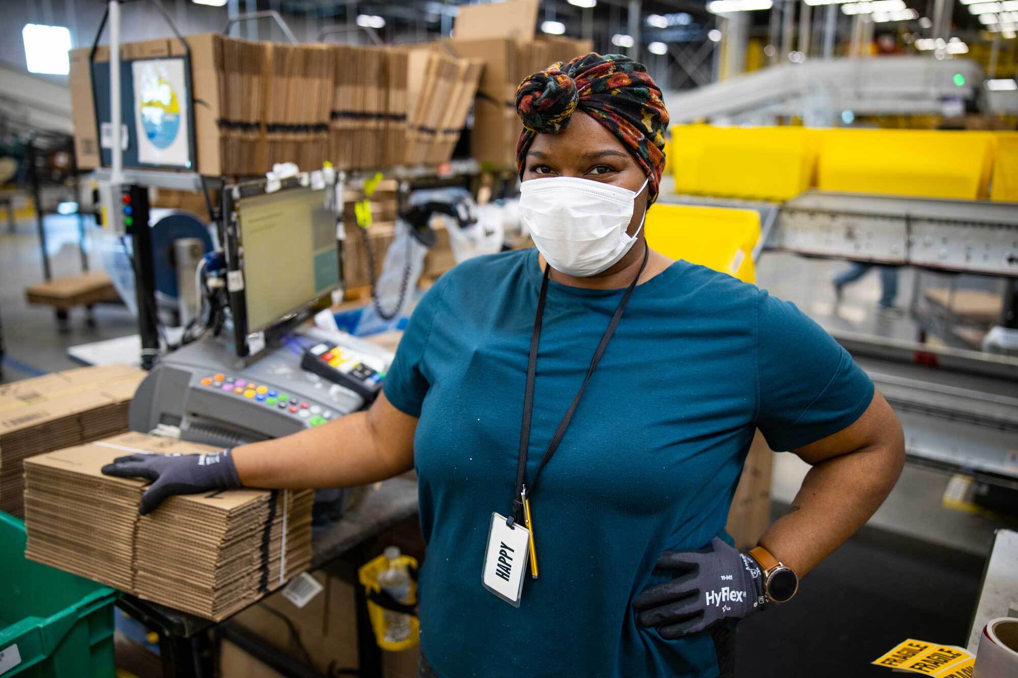 An Amazon associate working at a fulfillment center pack station poses for the camera while wearing a protective surgical mask