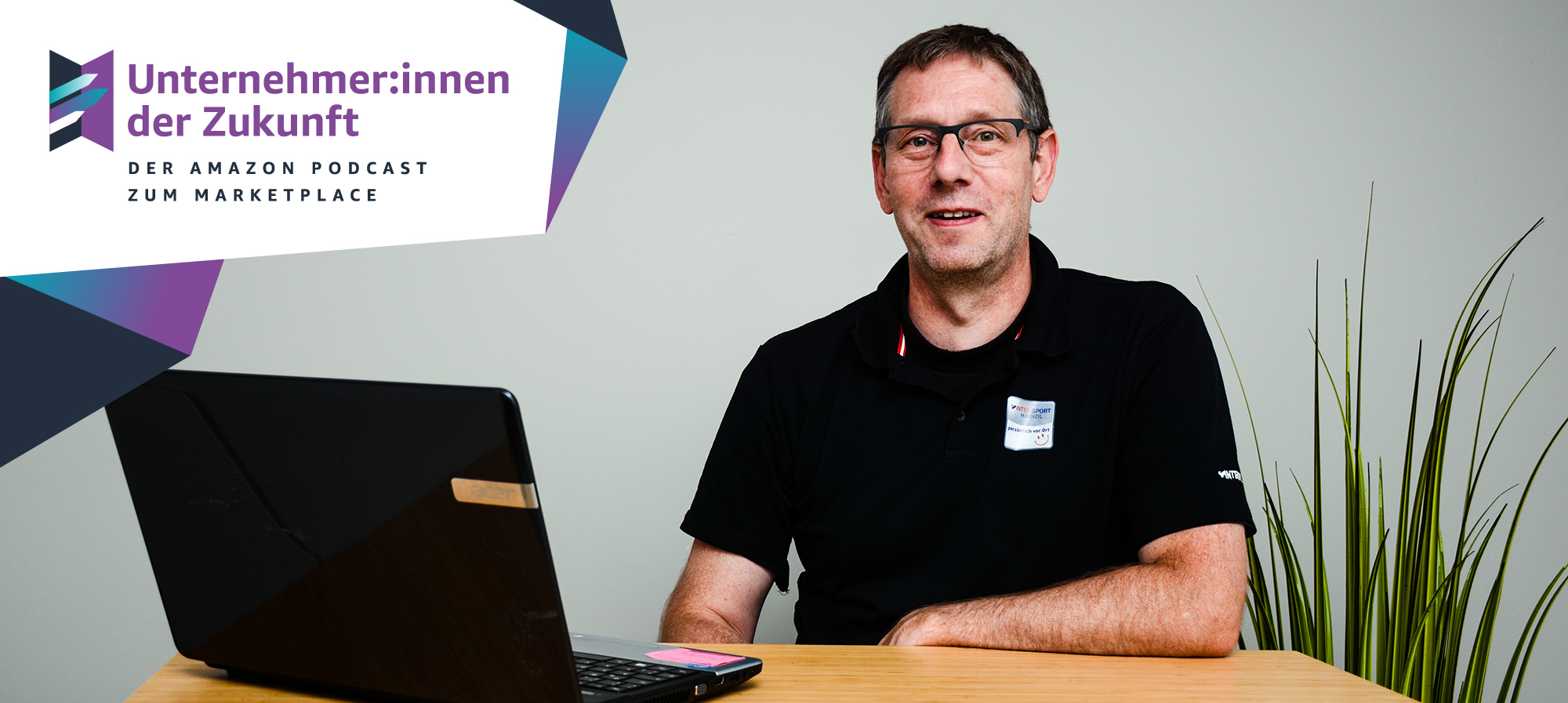 Ein Mann mit schwarzen Haaren und einem schwarzen T-shirt sitzt an einem Tisch. Vor ihm steht ein schwarzer Laptop.