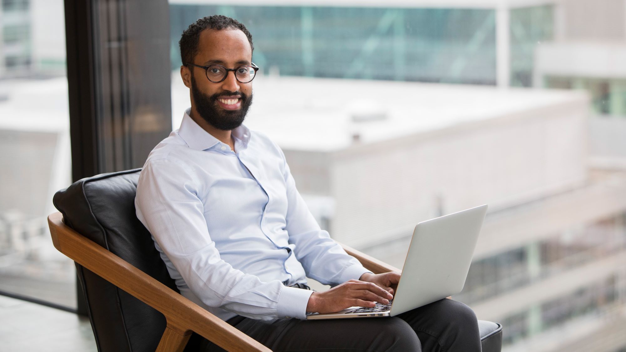 An image of a man seated in front of an office building window with a laptop.