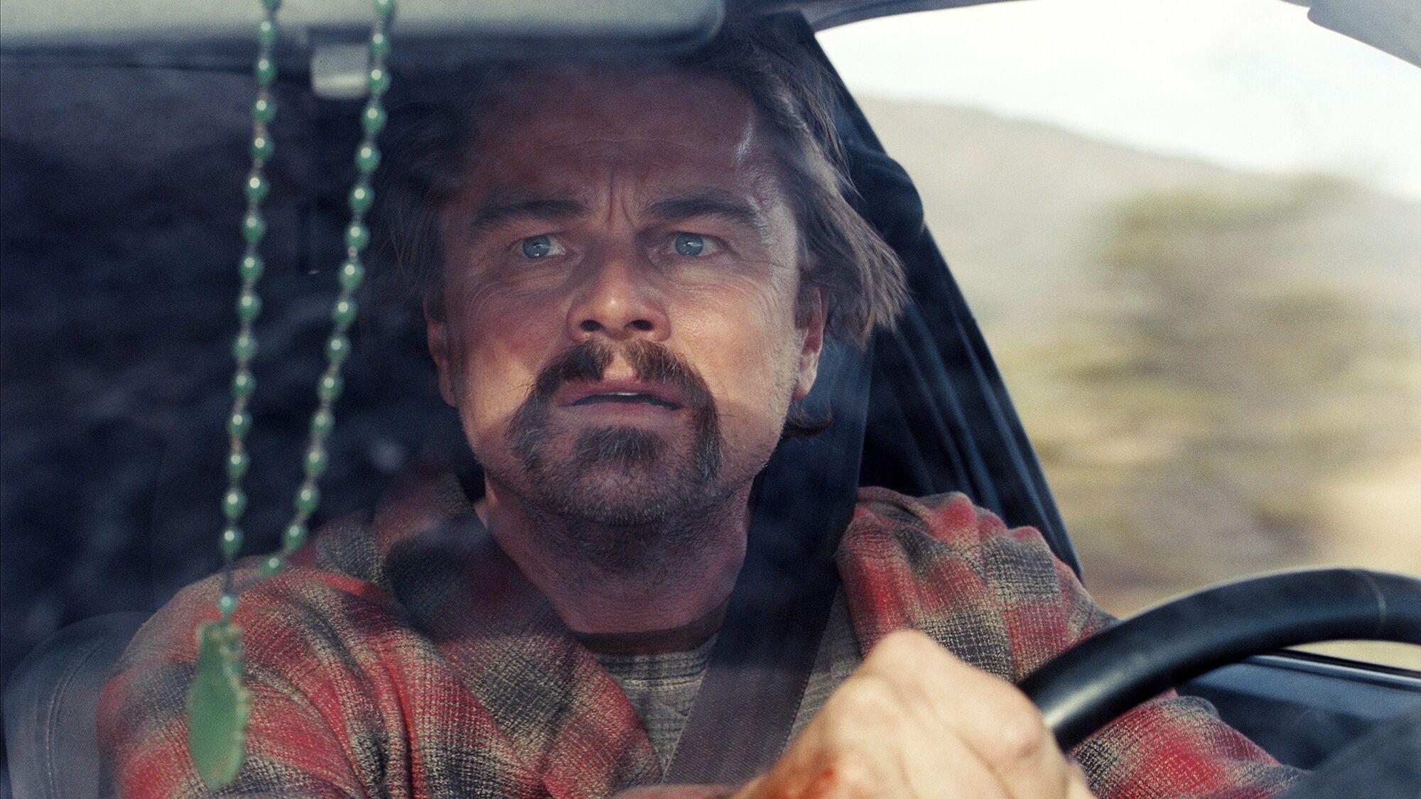 Bob Ferguson (Leonardo Di Caprio) with intense expression driving a car and wearing a plaid shirt.
