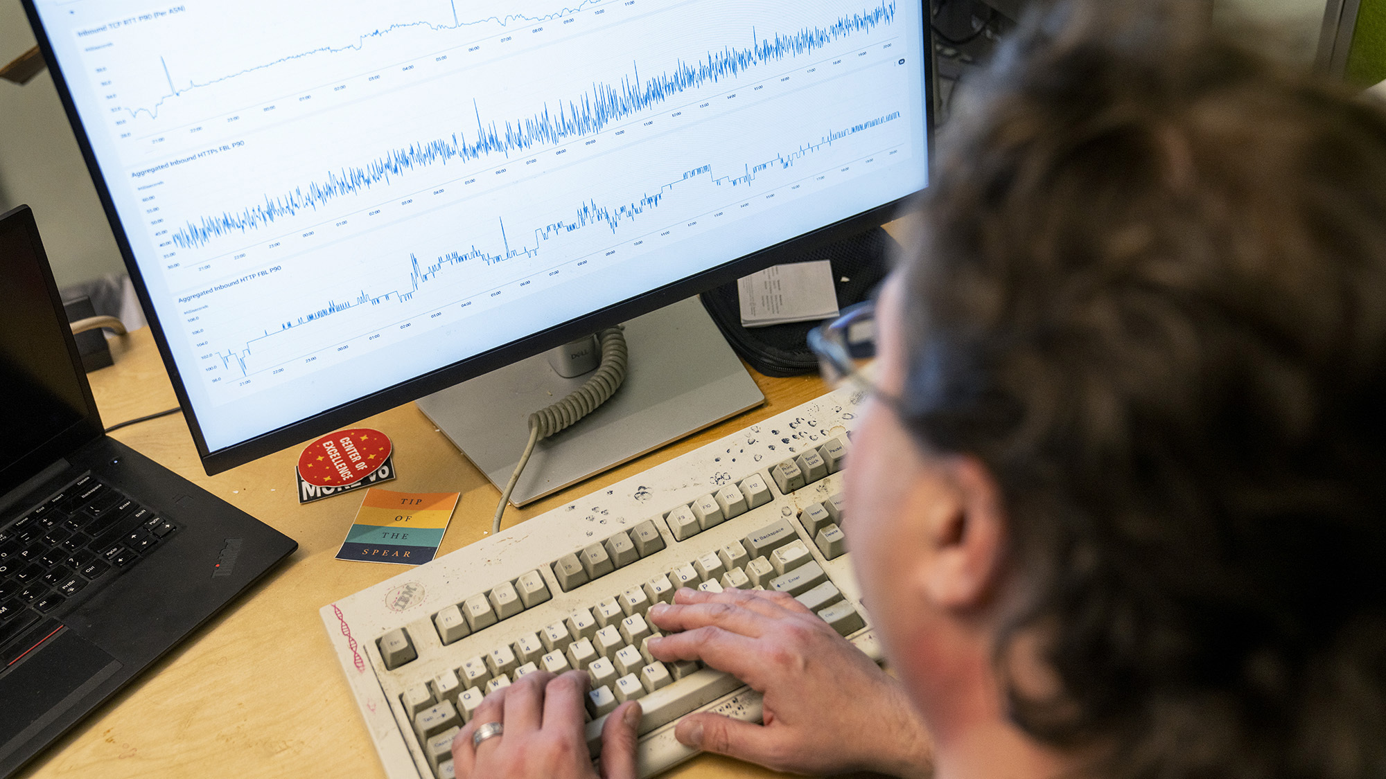 A photo of the back of Tom Scholl, AWS VP and Distinguished Engineer, sitting at this desk and working on the computer. Displayed on the Monitor is displaying internet traffic spikes.