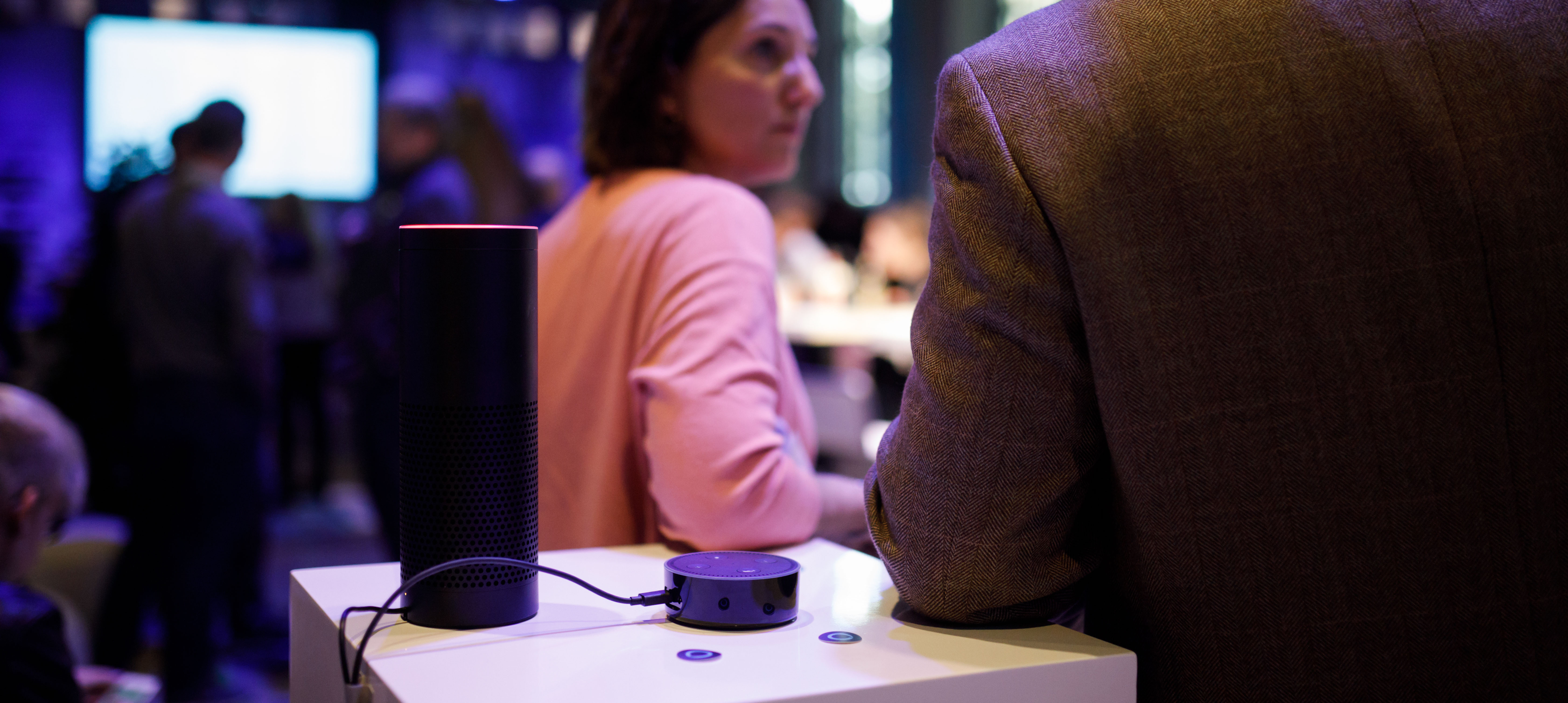 Ein Amazon Echo steht auf dem Tisch. Darum stehen Menschen, die sich unterhalten.